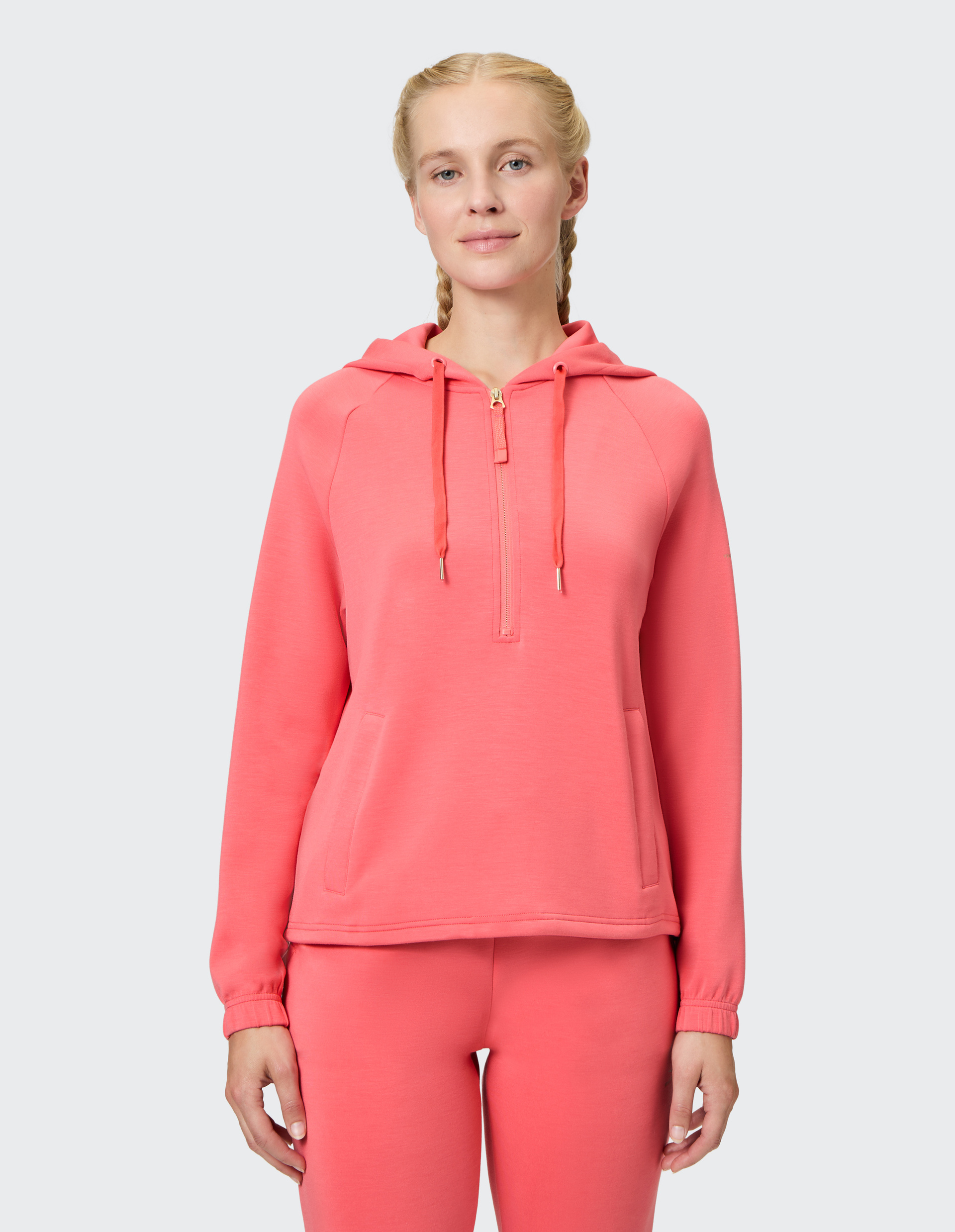 Frau mit geflochtenem Haar trägt rosa Kapuzenpulli und Hose vor hellem Hintergrund und blickt direkt in die Kamera.