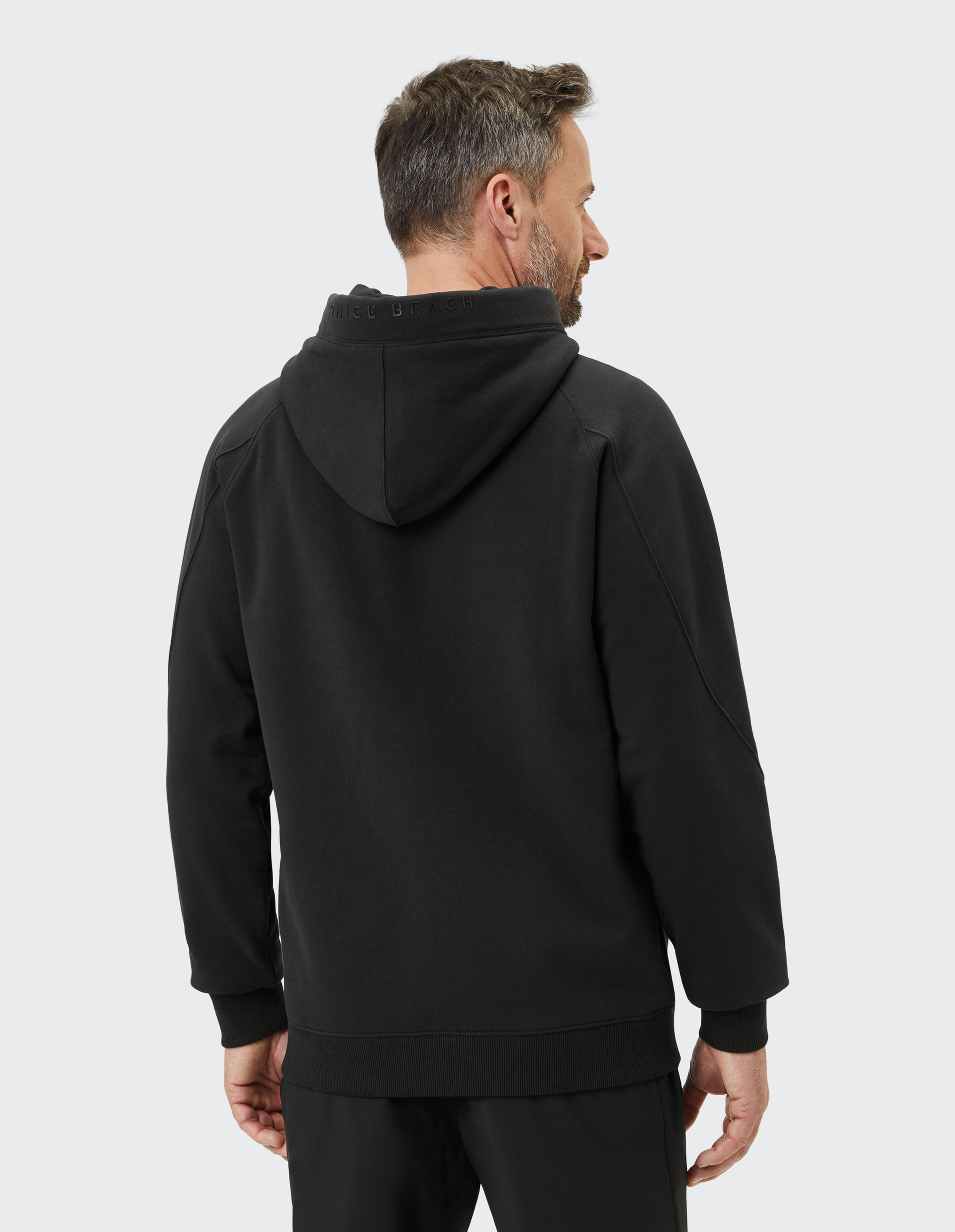 Ein Mann in schwarzem Kapuzenpullover steht mit dem Rücken zur Kamera, der Hintergrund ist einfarbig hellgrau.