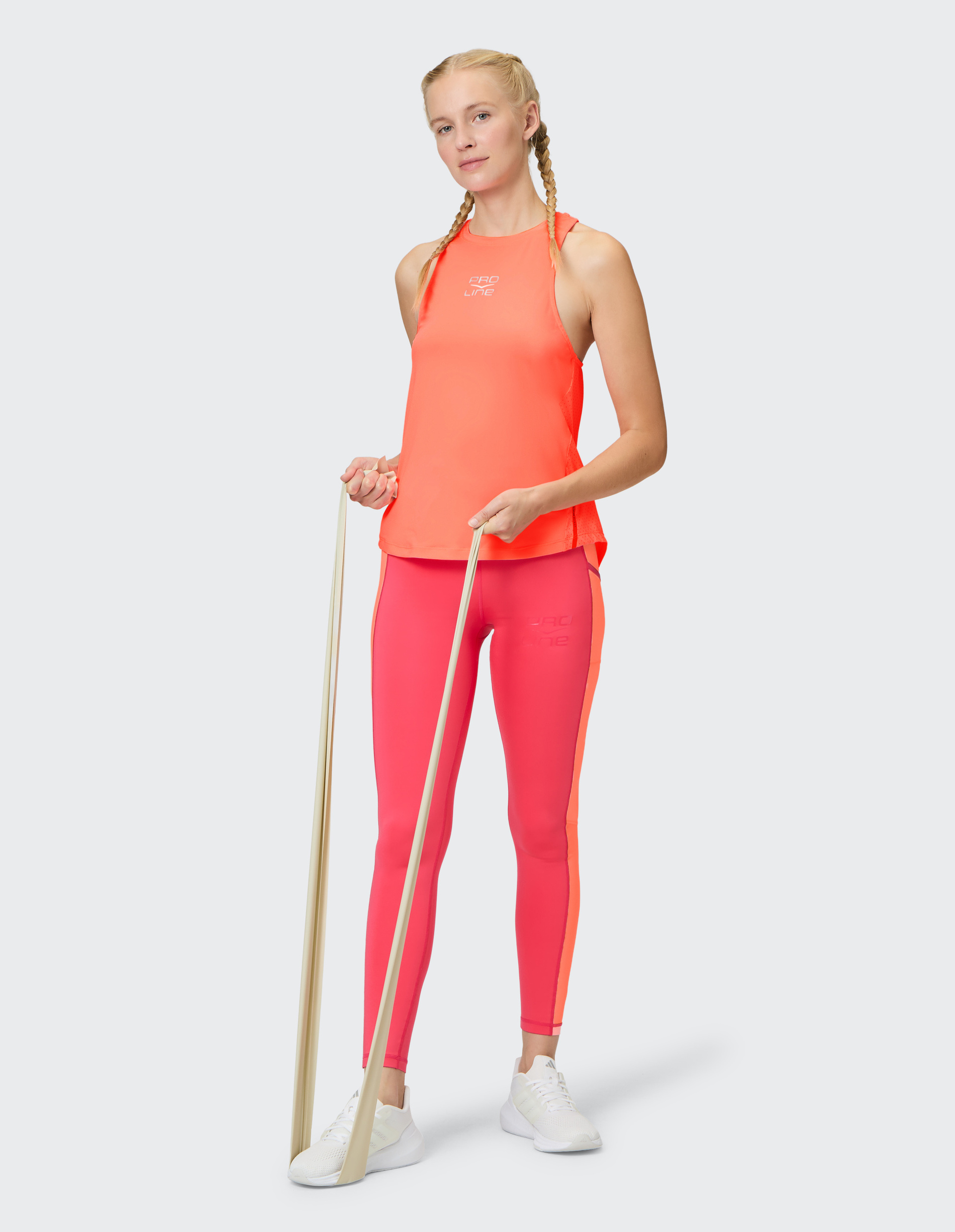 Frau in orange-rosa Sportkleidung mit Zopf hält ein Fitnessband, vor grauem Hintergrund, in weißem Schuhwerk stehend.