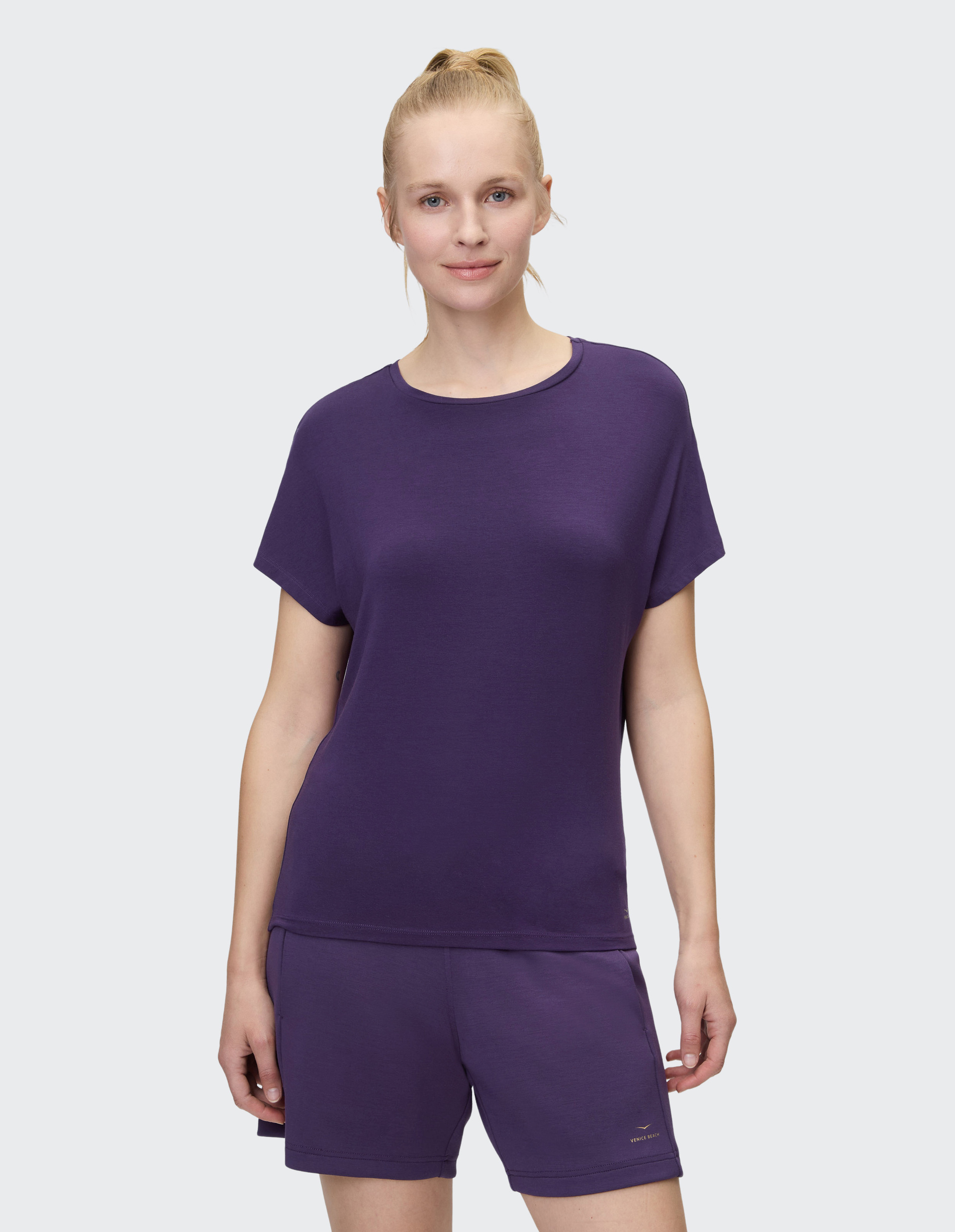 Eine Person trägt ein lila T-Shirt und passende Shorts, steht frontal und lächelt leicht vor neutralem Hintergrund.