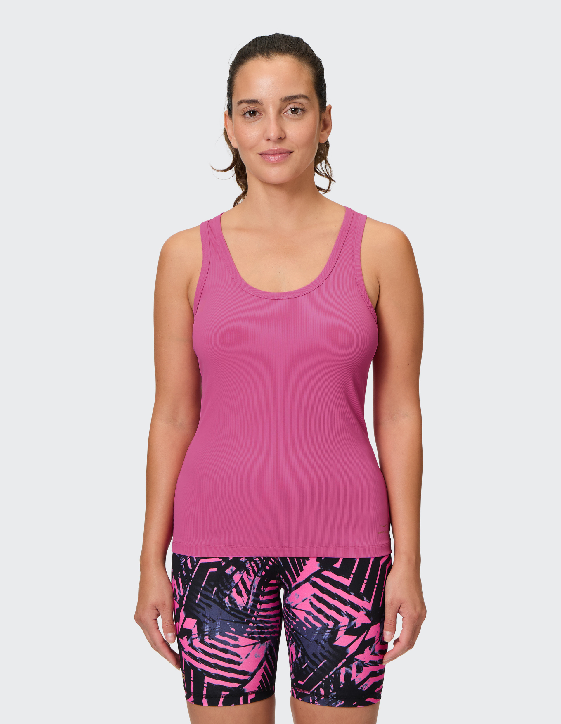 Frau in rosa Sporttop und gemusterten Shorts mit geometrischem Design steht vor neutralem Hintergrund und blickt freundlich zur Kamera.