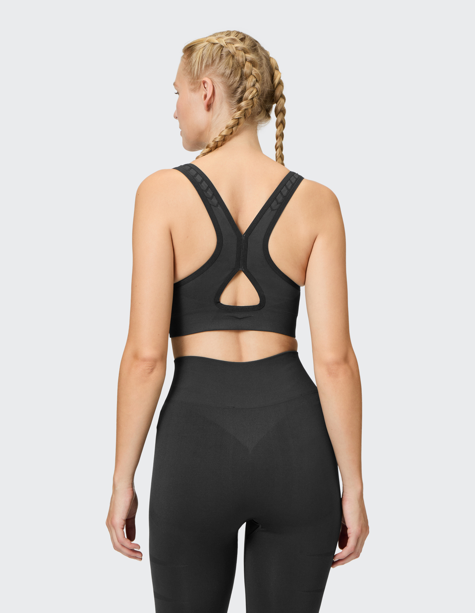 Frau mit geflochtenen Haaren trägt ein schwarzes Sportoutfit, bestehend aus Sport-BH und Leggings, zeigt den Rücken in Nahaufnahme.