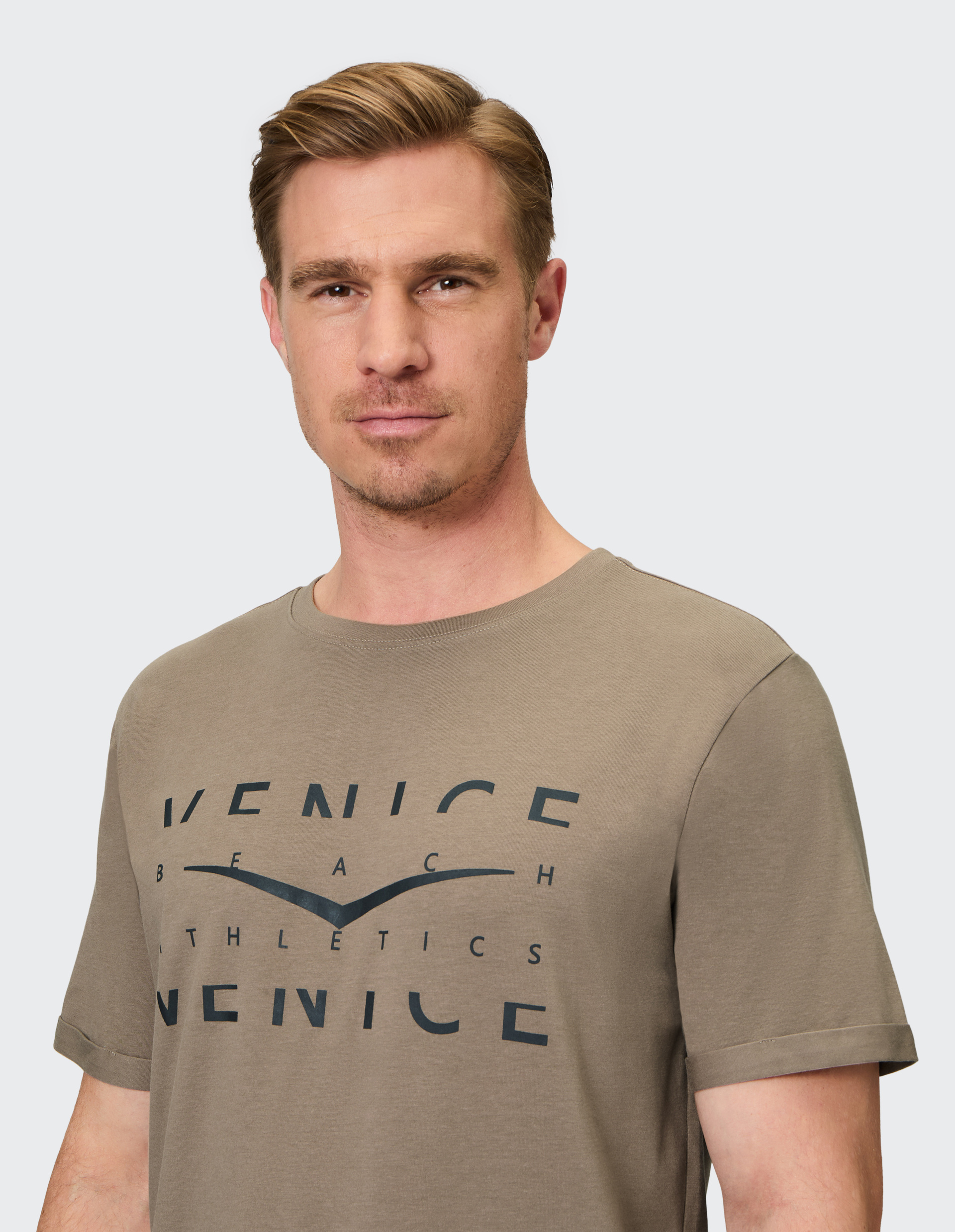 Mann mit kurzem Haar trägt ein olivgrünes T-Shirt mit Aufdruck "VENICE BEACH ATHLETICS" auf hellem Hintergrund.