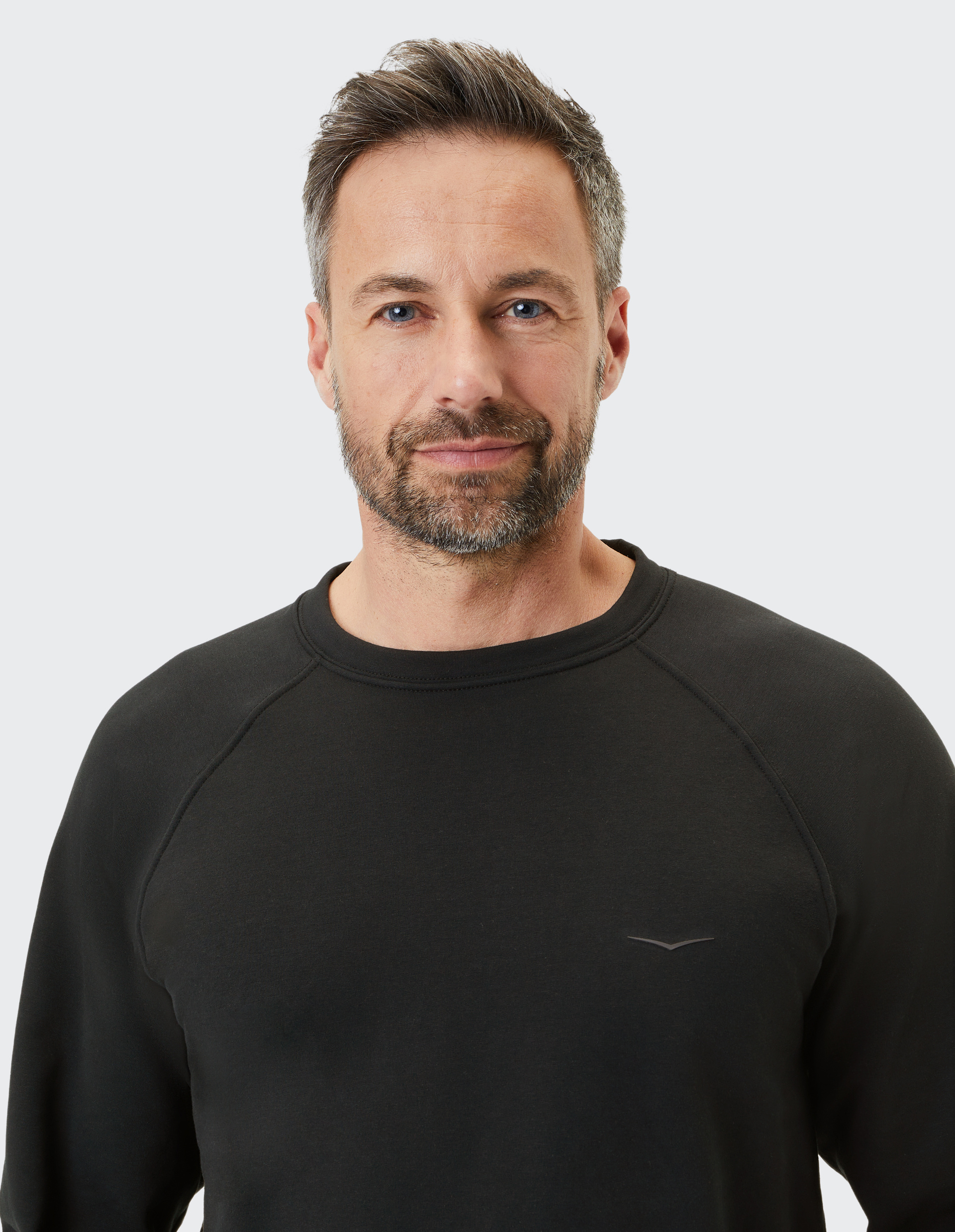 Ein lächelnder Mann mit Bart in einem schwarzen Pullover vor neutralem Hintergrund.