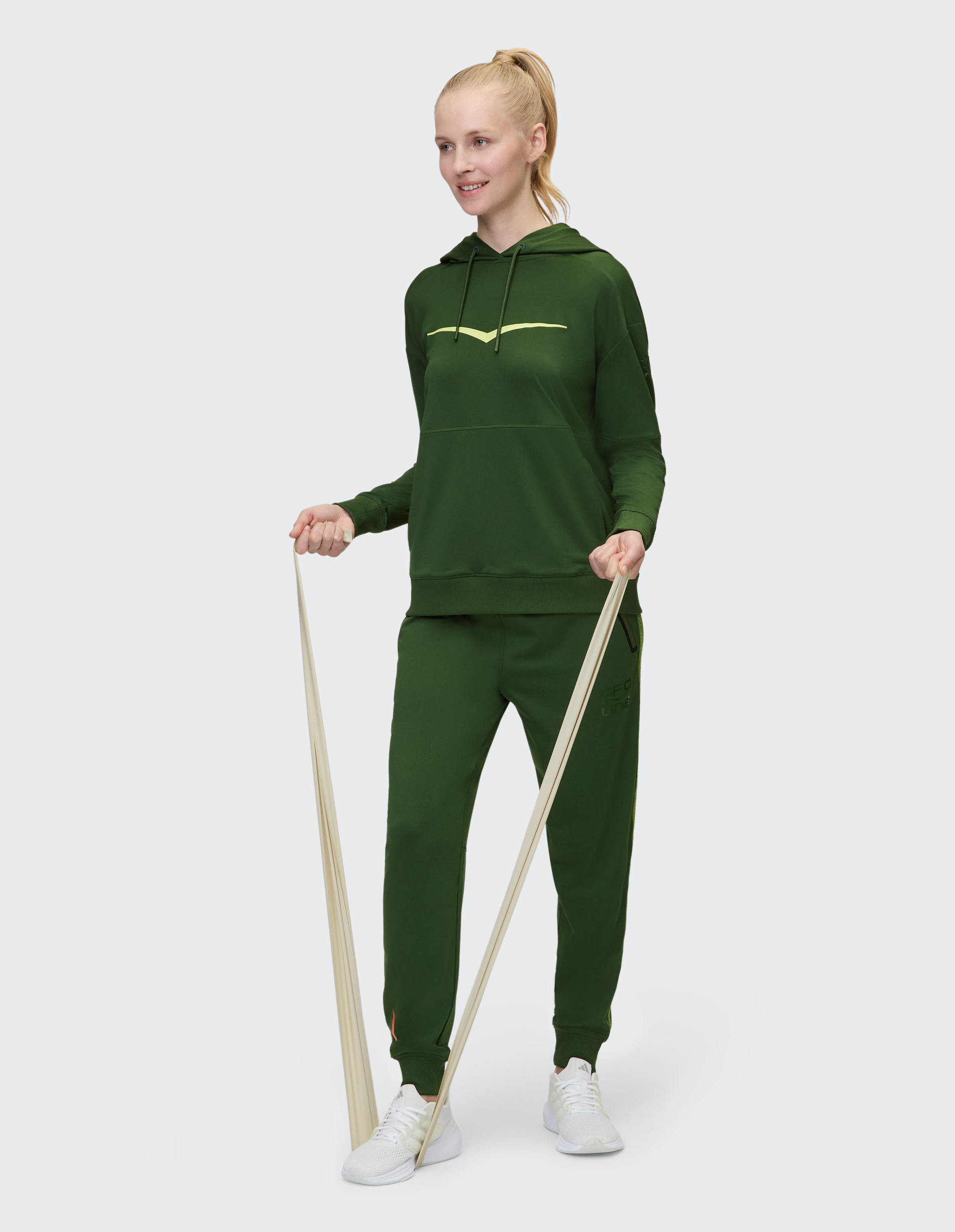 Frau in dunkelgrünem Sportanzug trainiert mit einem Fitnessband, lächelnd und in weißen Turnschuhen vor hellem Hintergrund.
