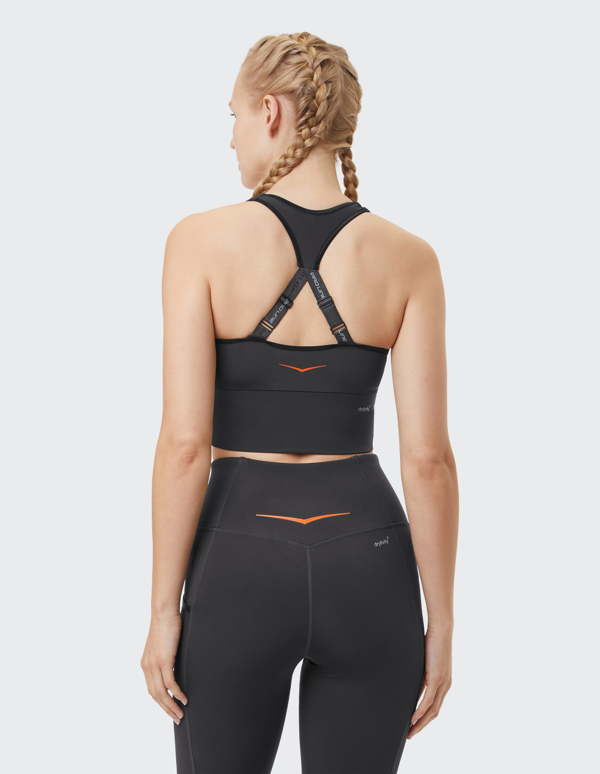 Frau in schwarzen Sportleggings und Top mit Kreuzträgern von hinten, blondes Haar in Zöpfen, steht vor neutralem Hintergrund.