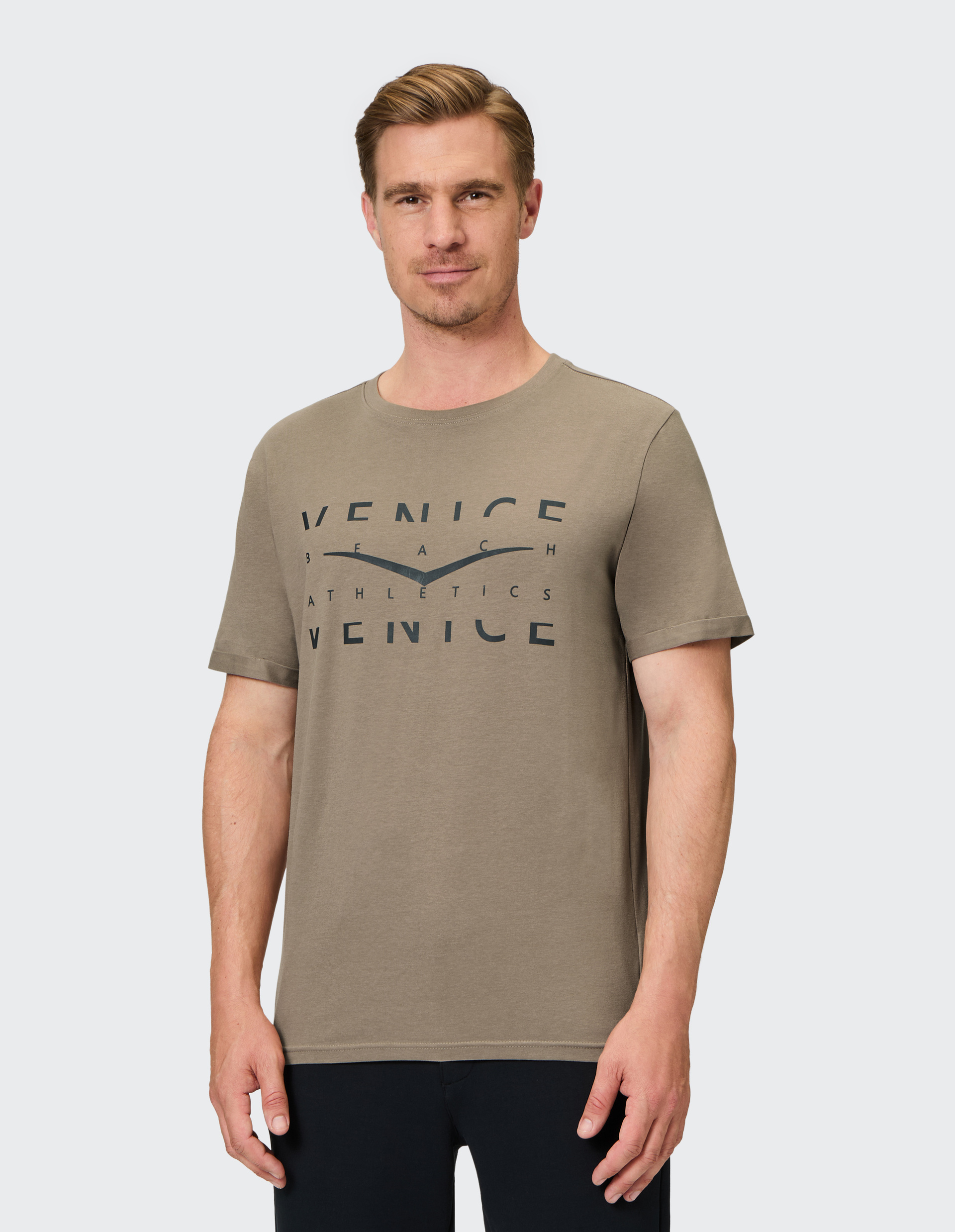 Mann in olivgrünem T-Shirt mit "Venice Beach Athletics"-Schriftzug steht lächelnd vor hellem Hintergrund, trägt schwarze Hose.