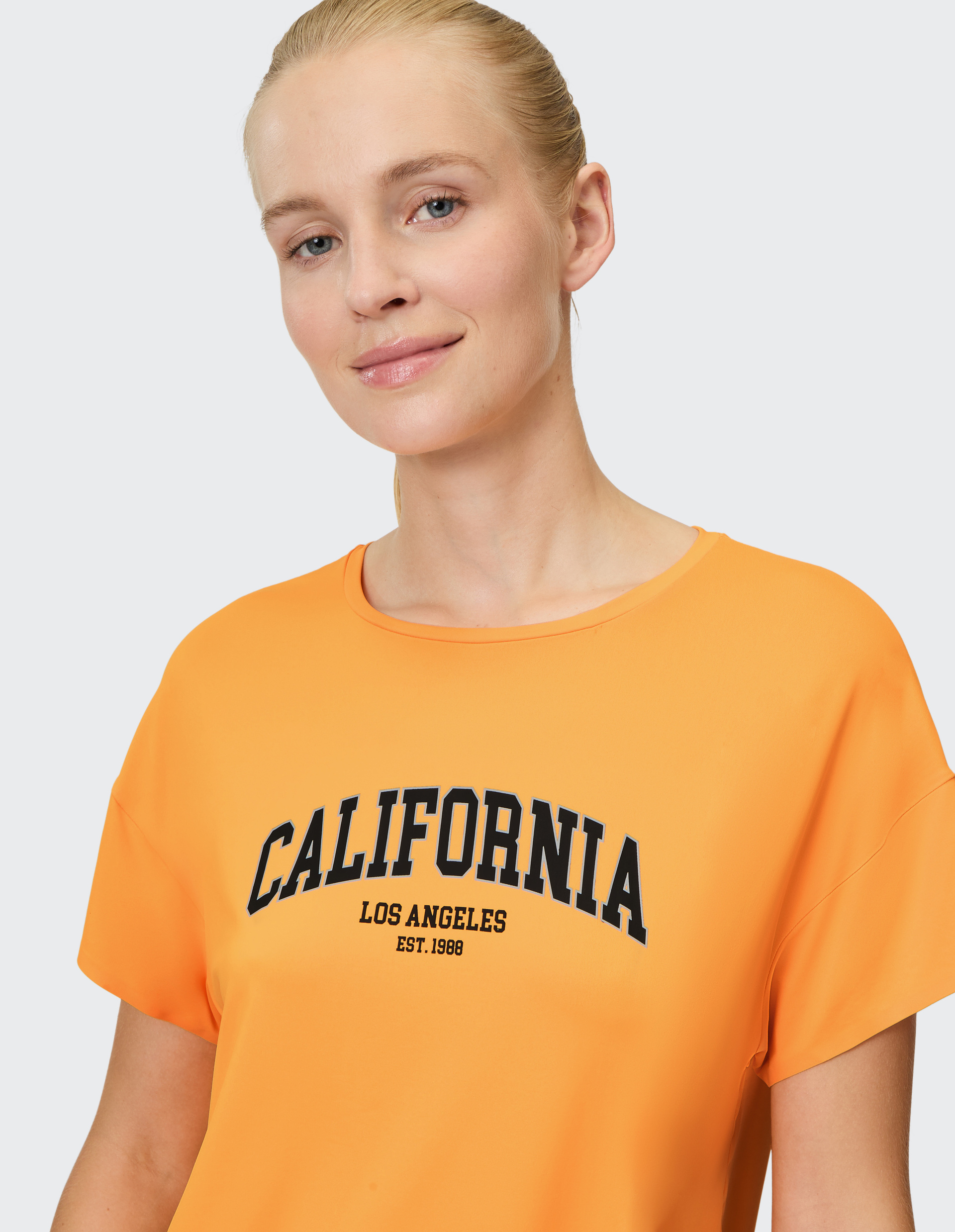 Frau in orangem T-Shirt mit Aufschrift "California Los Angeles" lächelt leicht, neutrale Hintergrundfarbe.