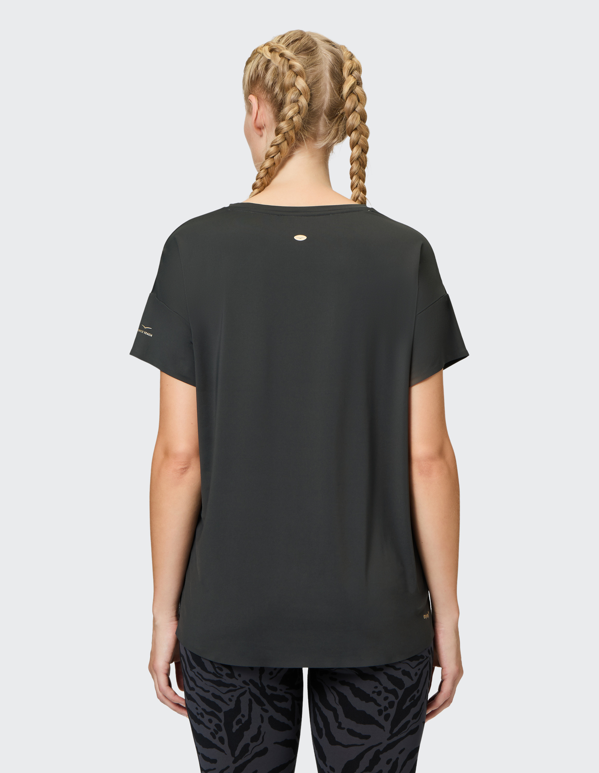 Eine Person mit zwei geflochtenen Zöpfen trägt ein schwarzes T-Shirt und gemusterte Leggings, von hinten fotografiert.