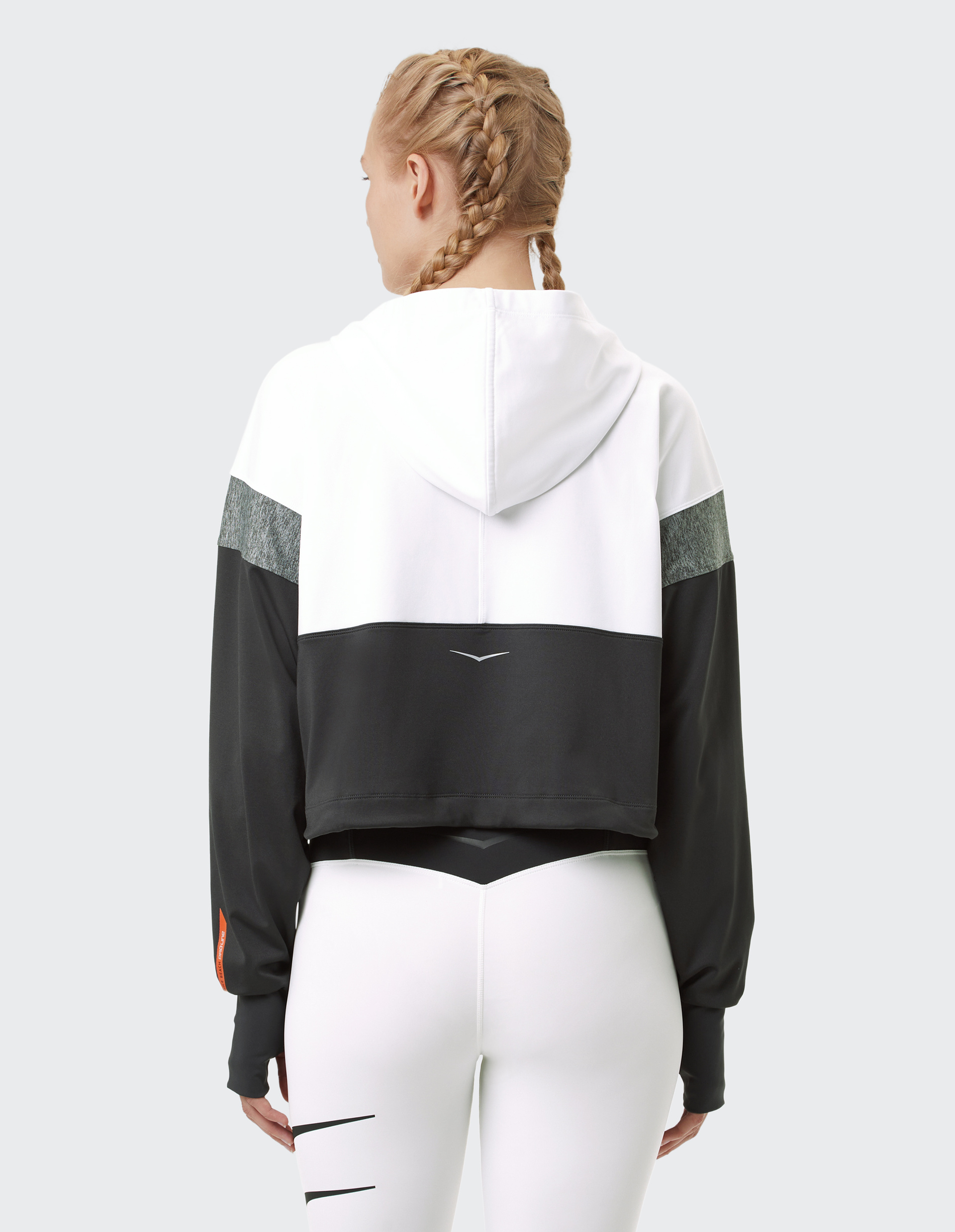 Frau mit geflochtenem Haar trägt einen zweifarbigen Hoodie und weiße Leggings, Rückenansicht vor grauem Hintergrund.