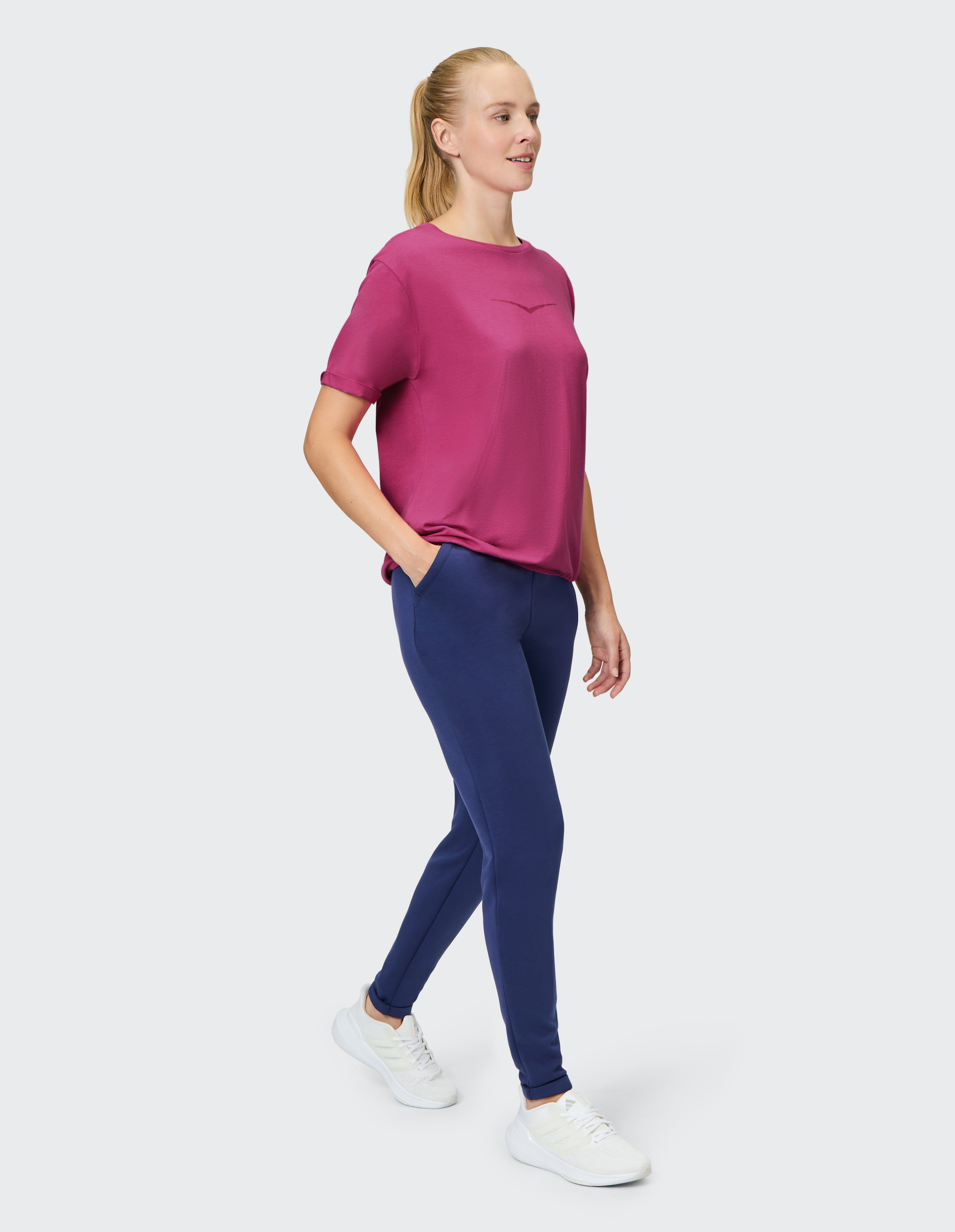 Frau in sportlicher Kleidung, pinkes T-Shirt und blaue Leggings, geht lächelnd mit Händen in den Taschen und trägt weiße Turnschuhe.