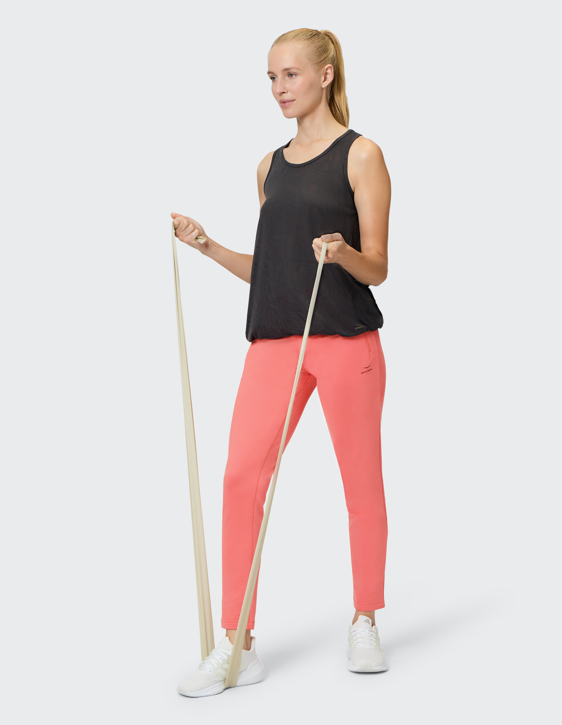 Frau in schwarzem Tanktop und korallfarbenen Leggings nutzt ein Fitnessband für Übungen, steht auf einem Bein, lächelt leicht.