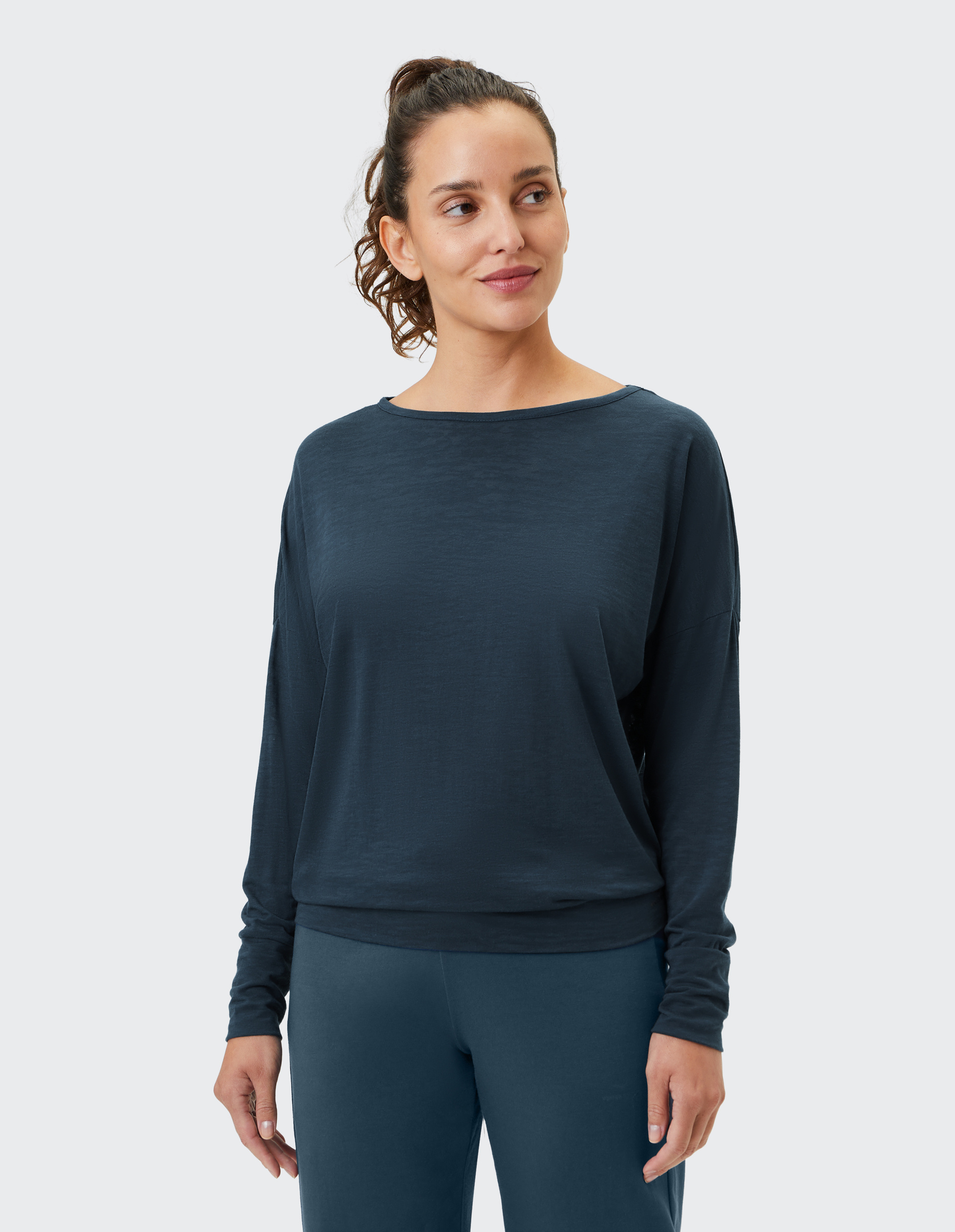 Frau in dunkelblauem Langarmshirt und passender Hose, lächelt leicht, mit lockigem Haar, vor hellem Hintergrund.