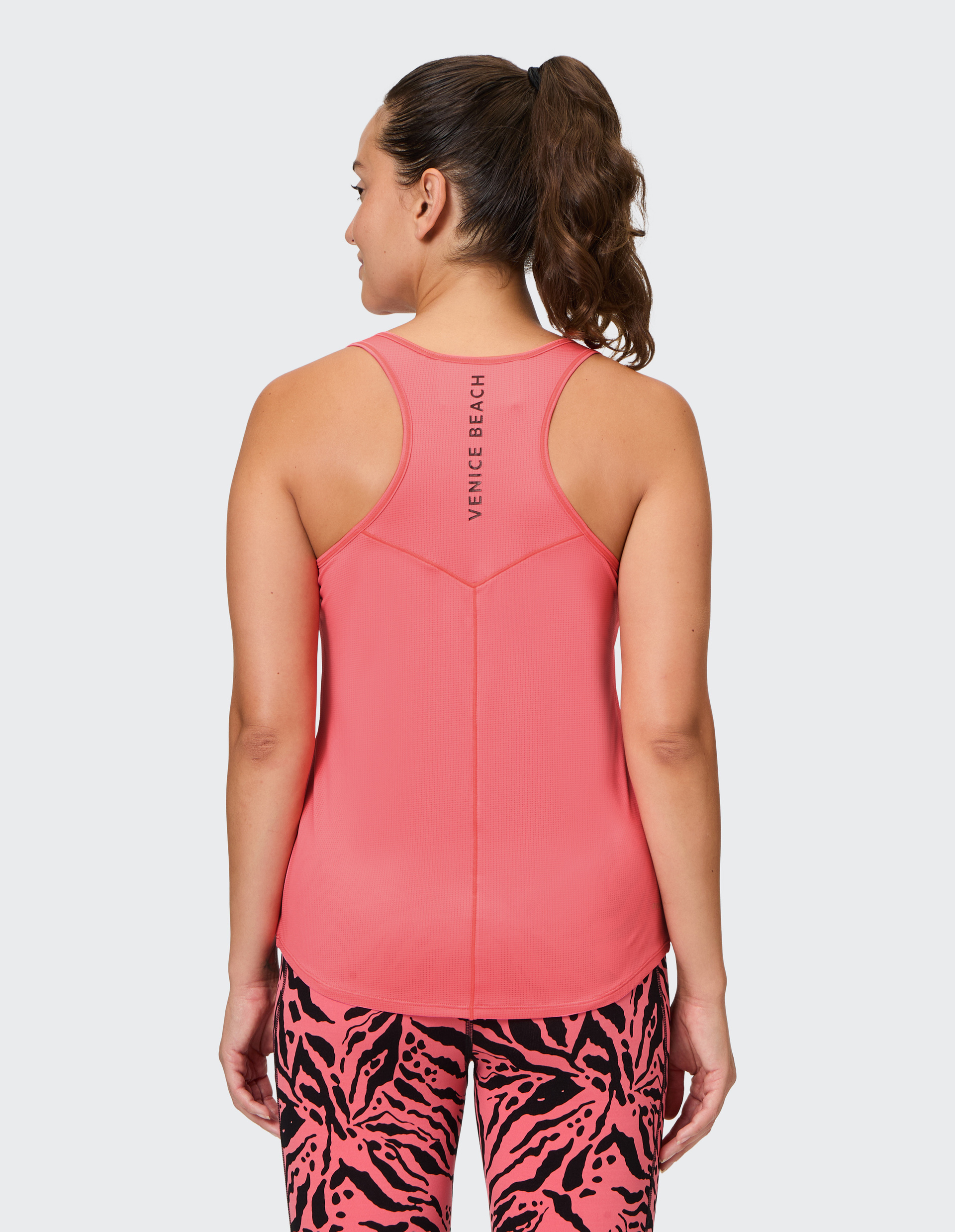 Eine Frau in einem pinken Sporttop mit "Venice Beach"-Schriftzug und gemusterten Leggings steht mit dem Rücken zur Kamera.