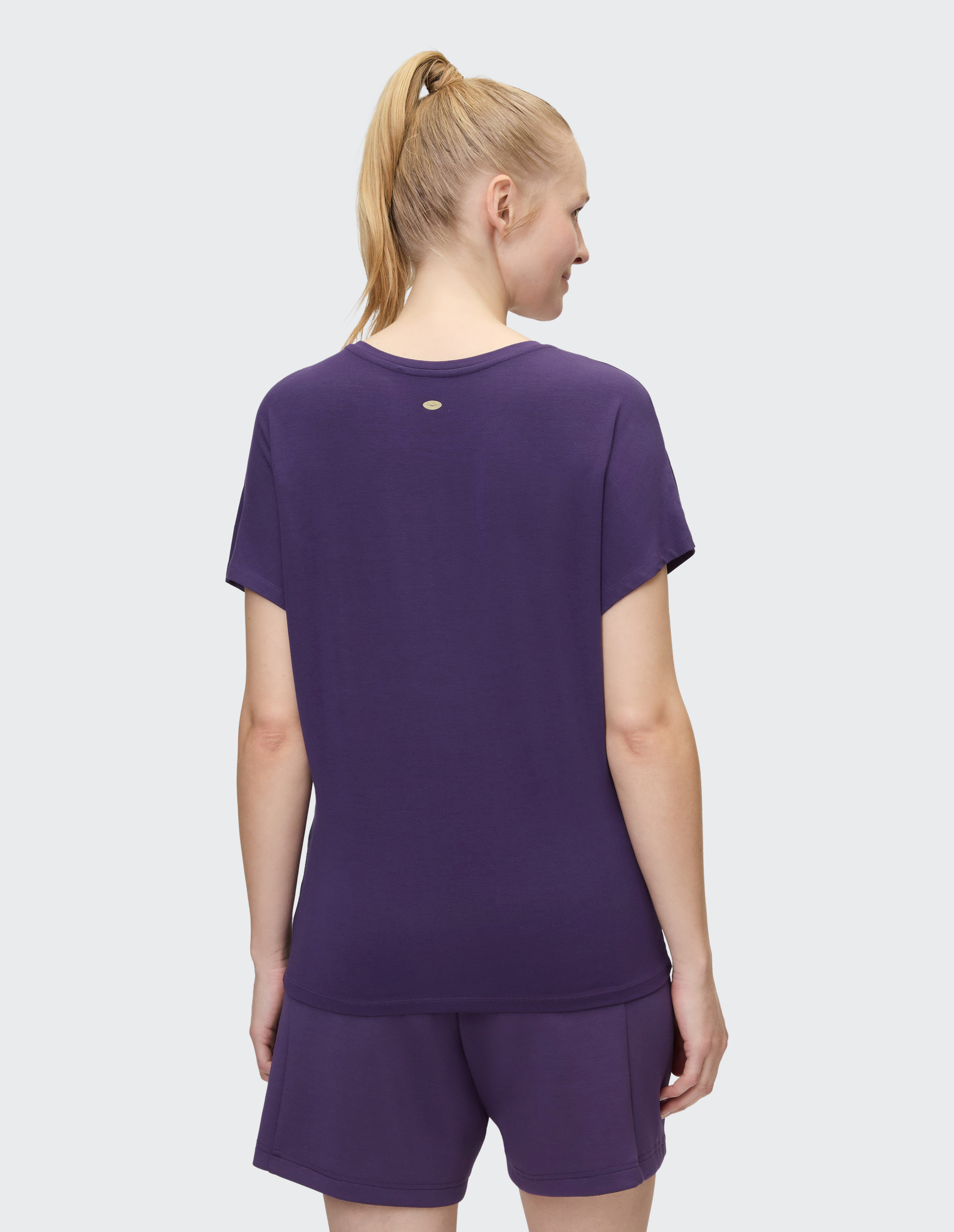 Frau von hinten in violettem T-Shirt und Shorts, mit blondem Pferdeschwanz vor grauem Hintergrund stehend.