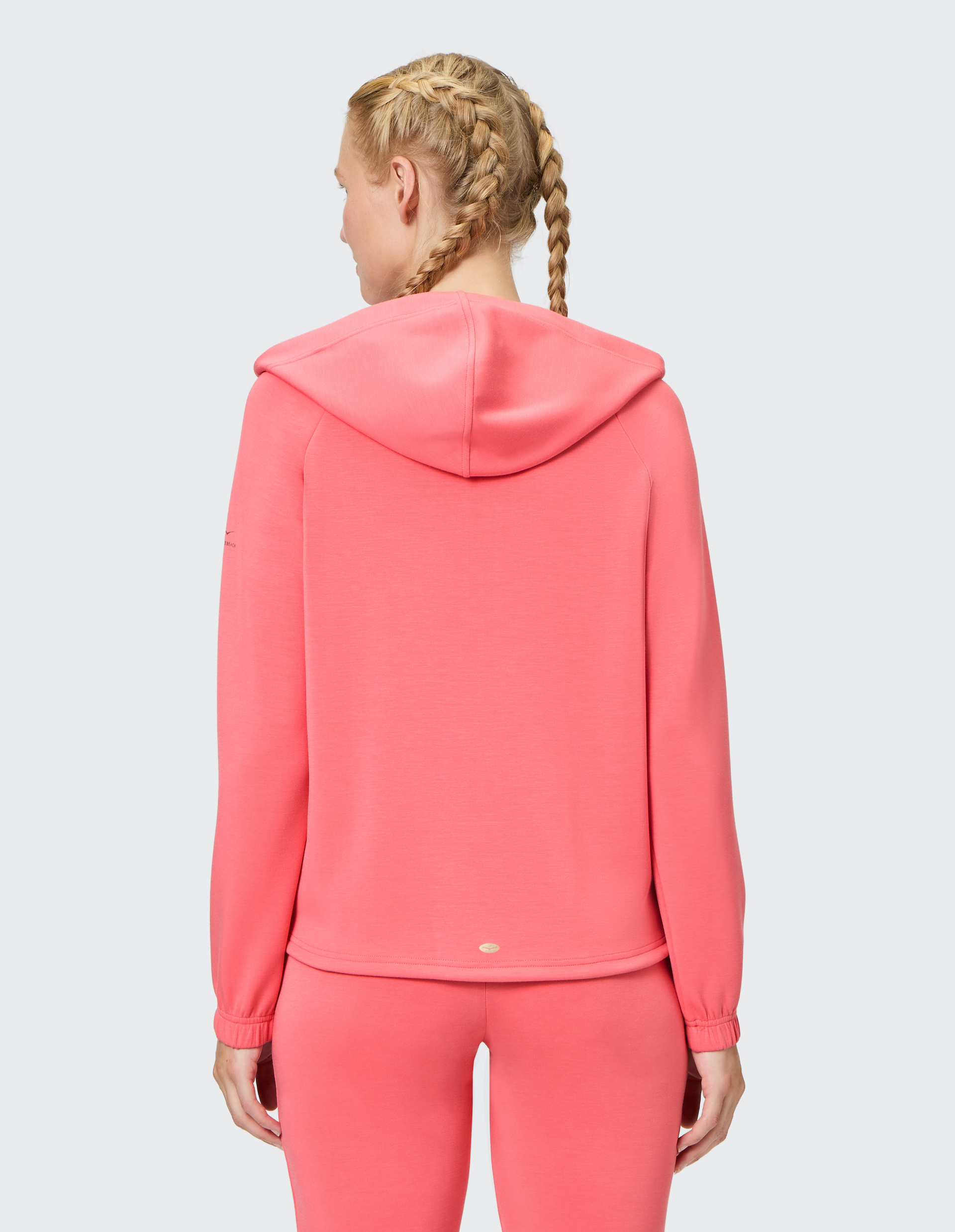 Person von hinten in rosa Kapuzenpullover und Hose, mit zwei geflochtenen Zöpfen, auf neutralem Hintergrund.