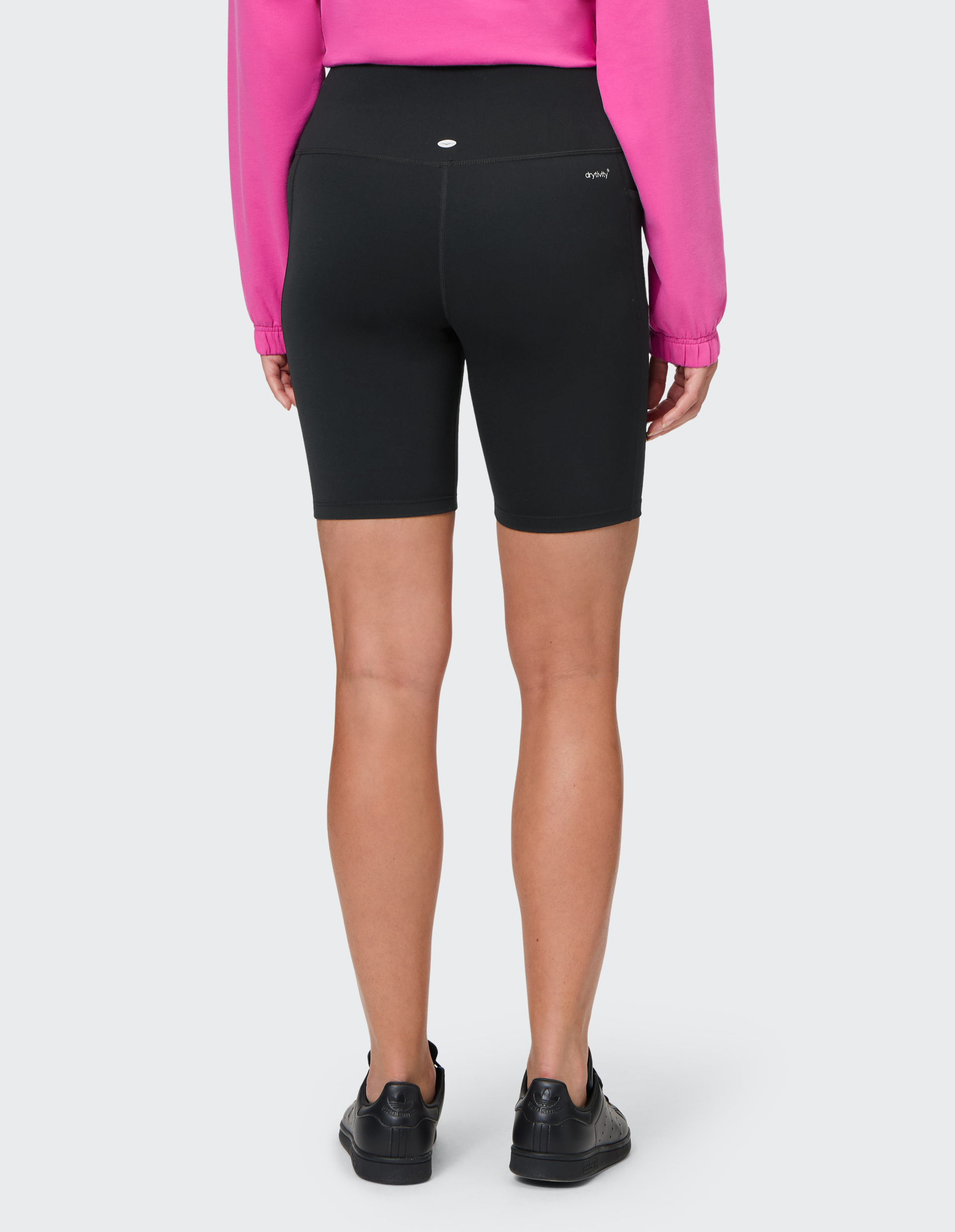 Person von hinten, trägt schwarzen Shorts, pinken Pullover und schwarze Sneaker, steht vor grauem Hintergrund.