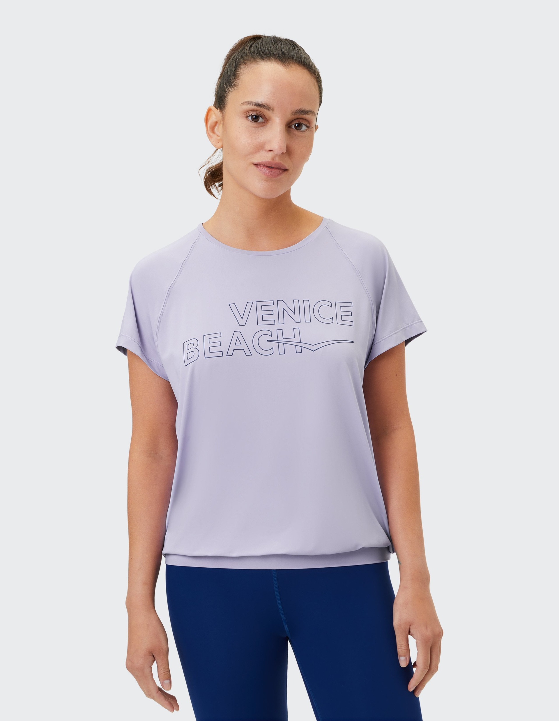 Eine Frau trägt ein hellviolettes T-Shirt mit "VENICE BEACH"-Aufdruck und dunkelblauen Leggings.