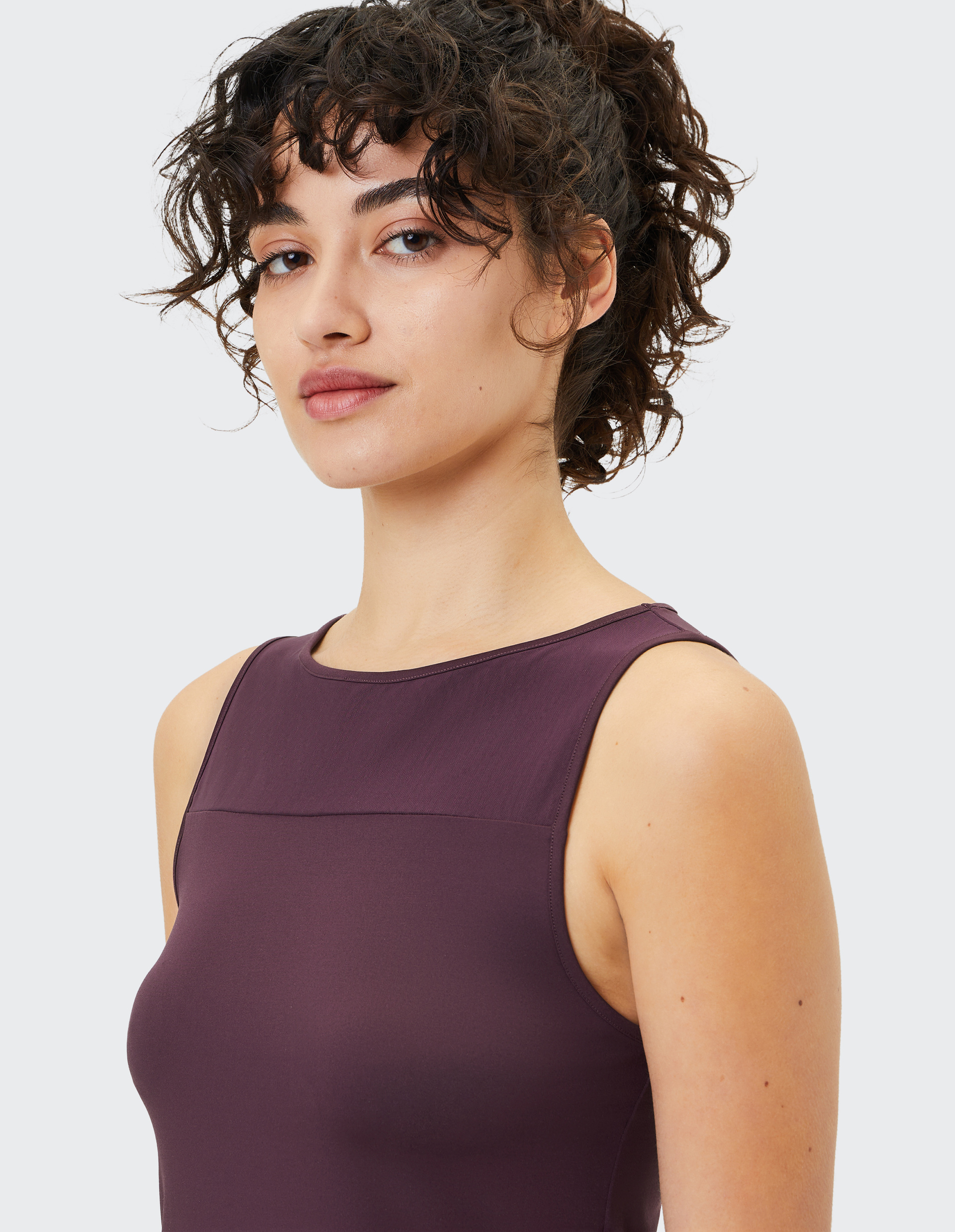 Frau mit lockigem Haar trägt ein ärmelloses, dunkelviolettes Oberteil und blickt mit einem leichten Lächeln zur Seite.