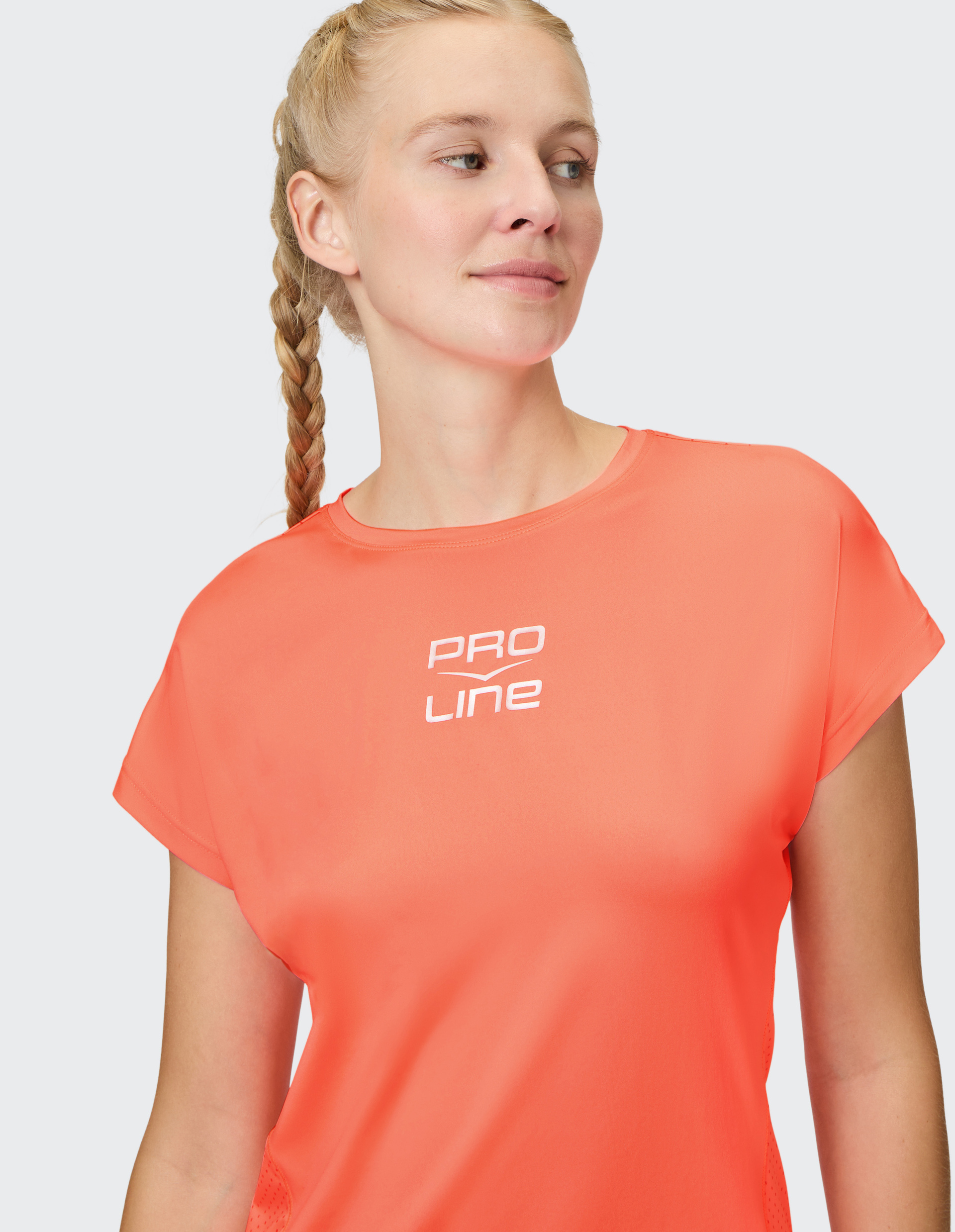 Frau in korallefarbenem Pro Line T-Shirt, guckt zur Seite, trägt einen geflochtenen Zopf; vor grauem Hintergrund.