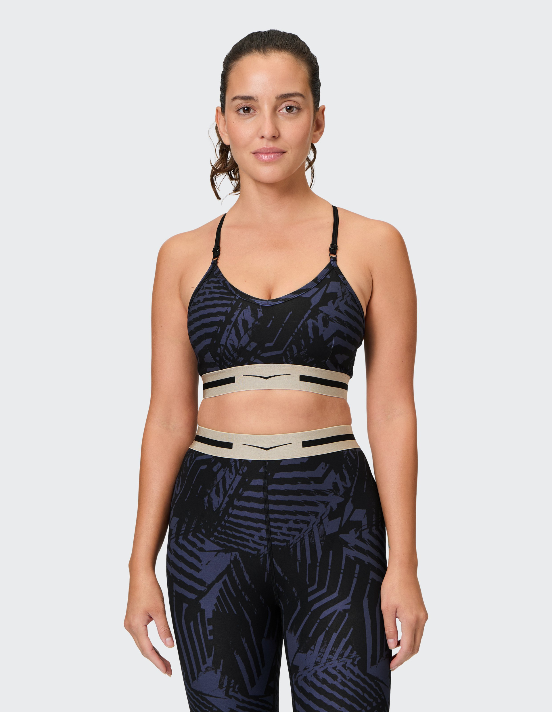 Frau in einem gemusterten Sport-BH und Leggings posiert vor einem neutralen Hintergrund, lächelt mit locker zurückgebundenem Haar.