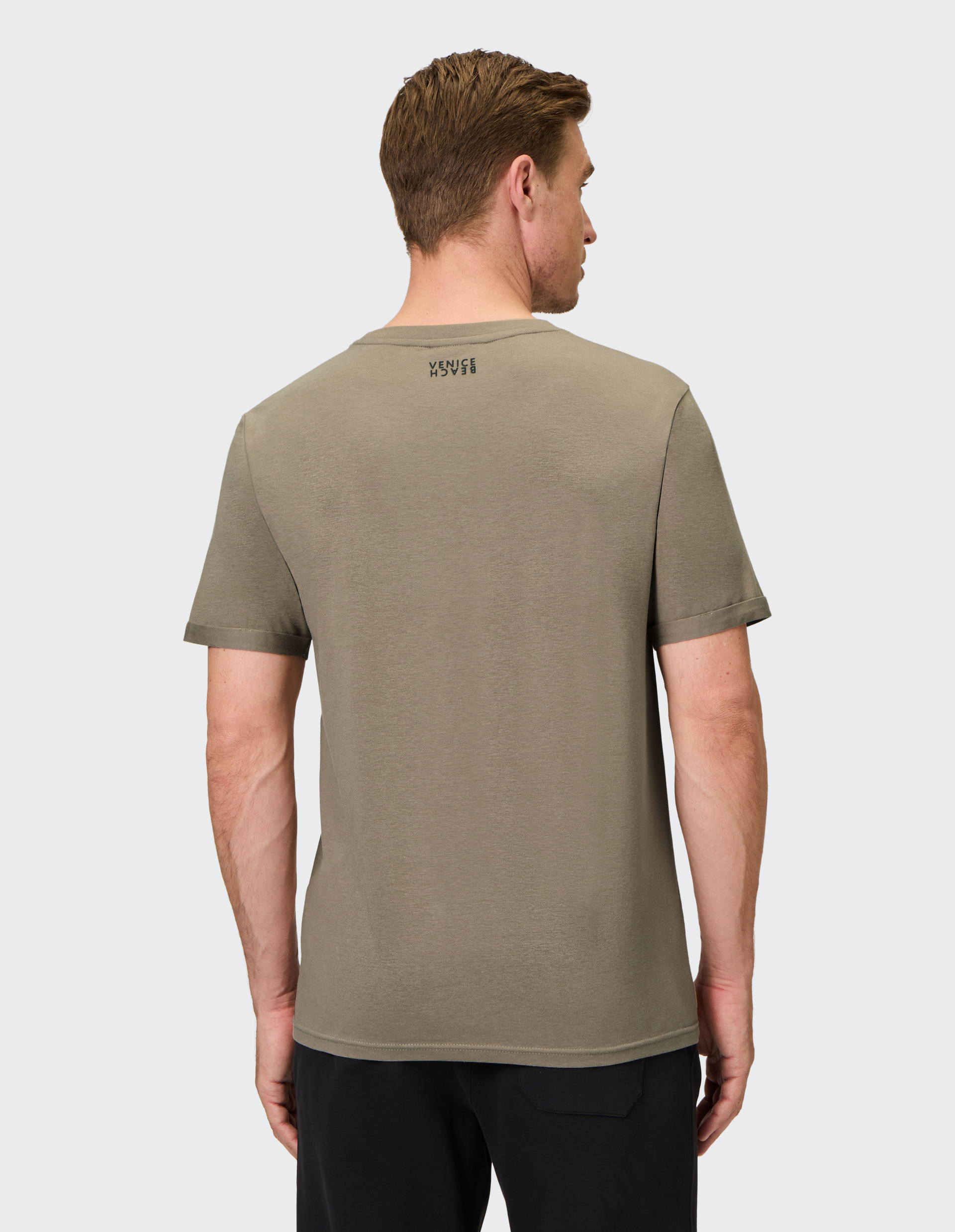 Ein Mann von hinten in einem olivgrünen T-Shirt mit schwarzem Logo am oberen Rücken und schwarzen Hosen, auf grauem Hintergrund.