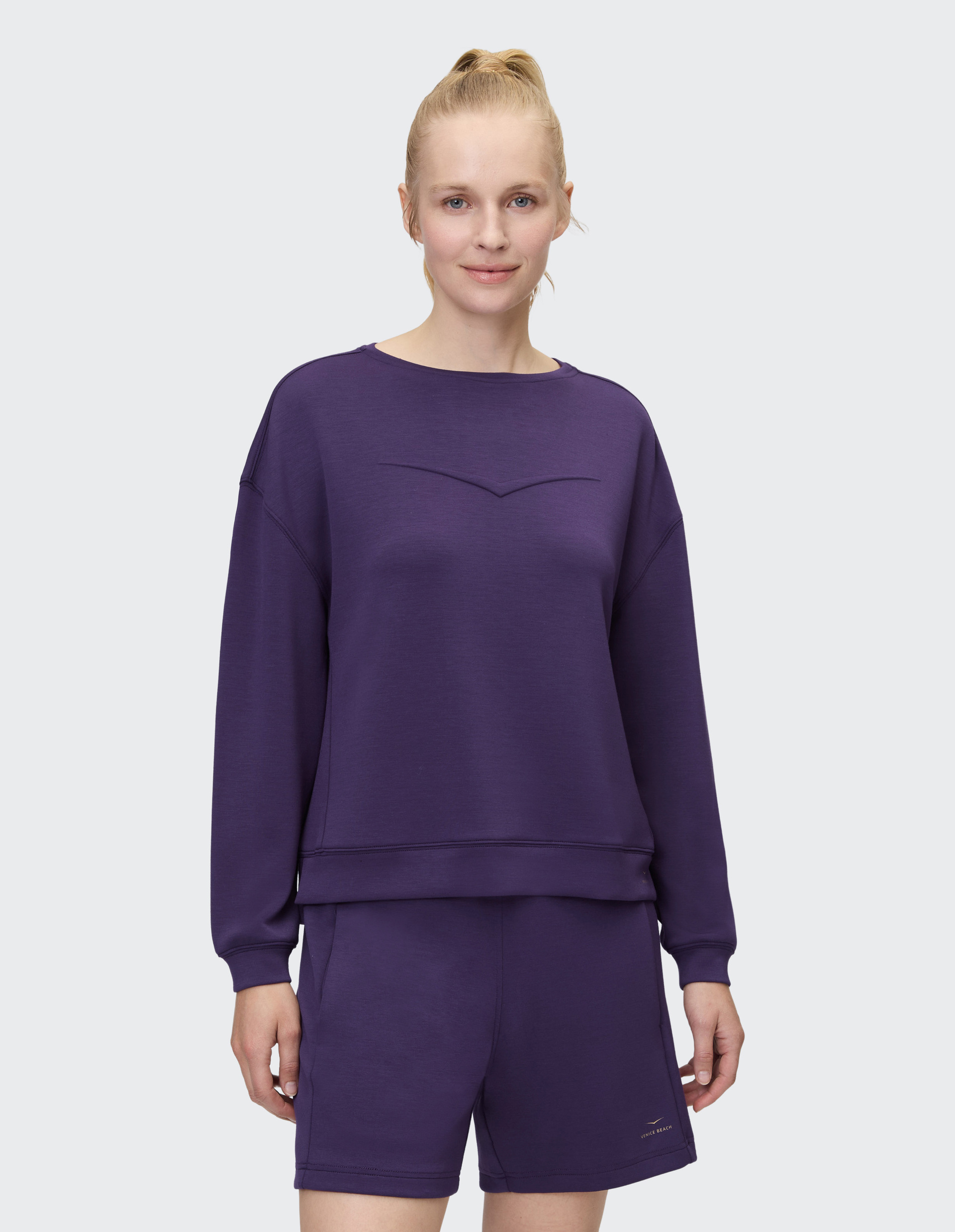 Eine lächelnde Person in lila Sweatshirt und Shorts steht vor einem grauen Hintergrund, Haare im Pferdeschwanz gebunden.
