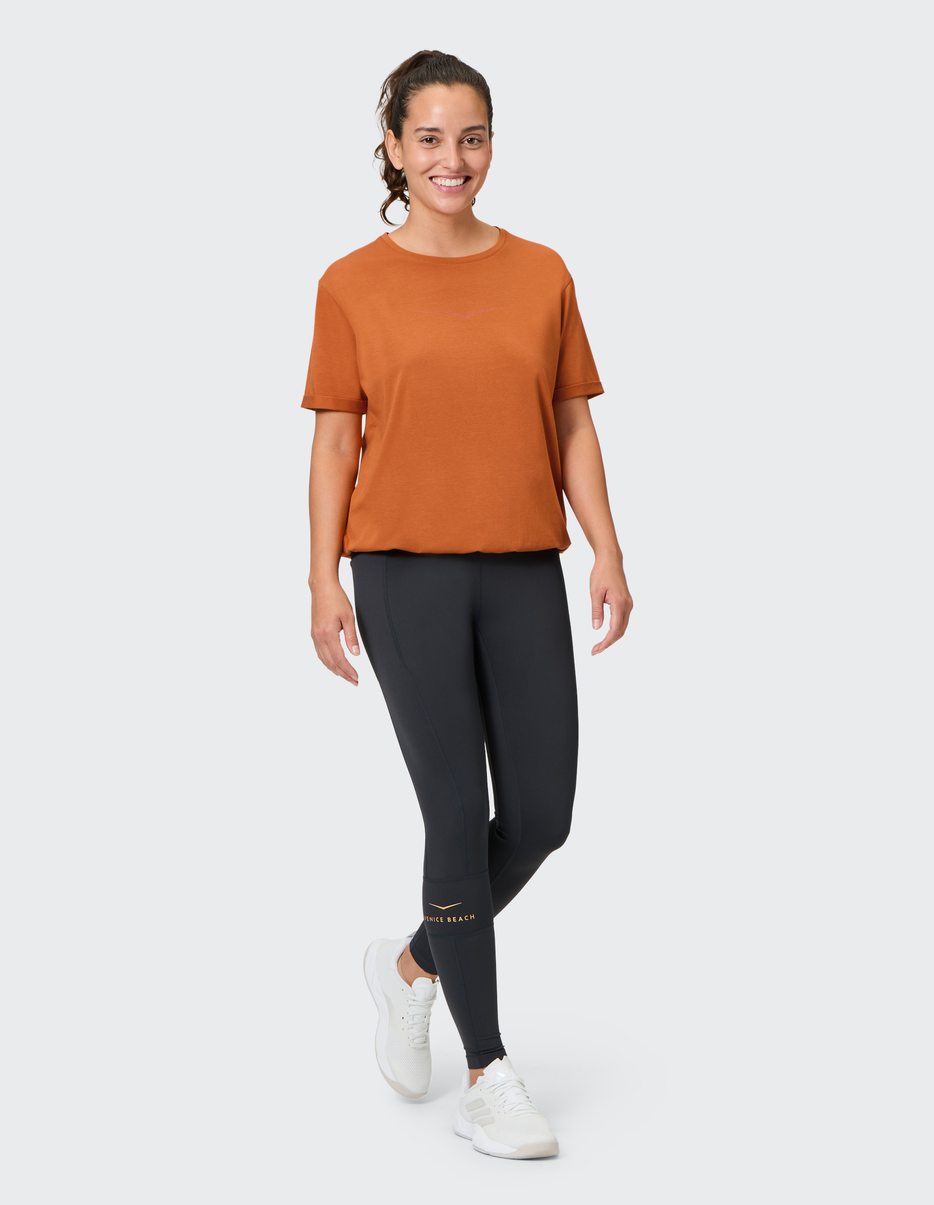 Frau in orangefarbenem T-Shirt und dunkelgrauer Leggings mit weißen Sneakers lächelt und steht vor neutralem Hintergrund.