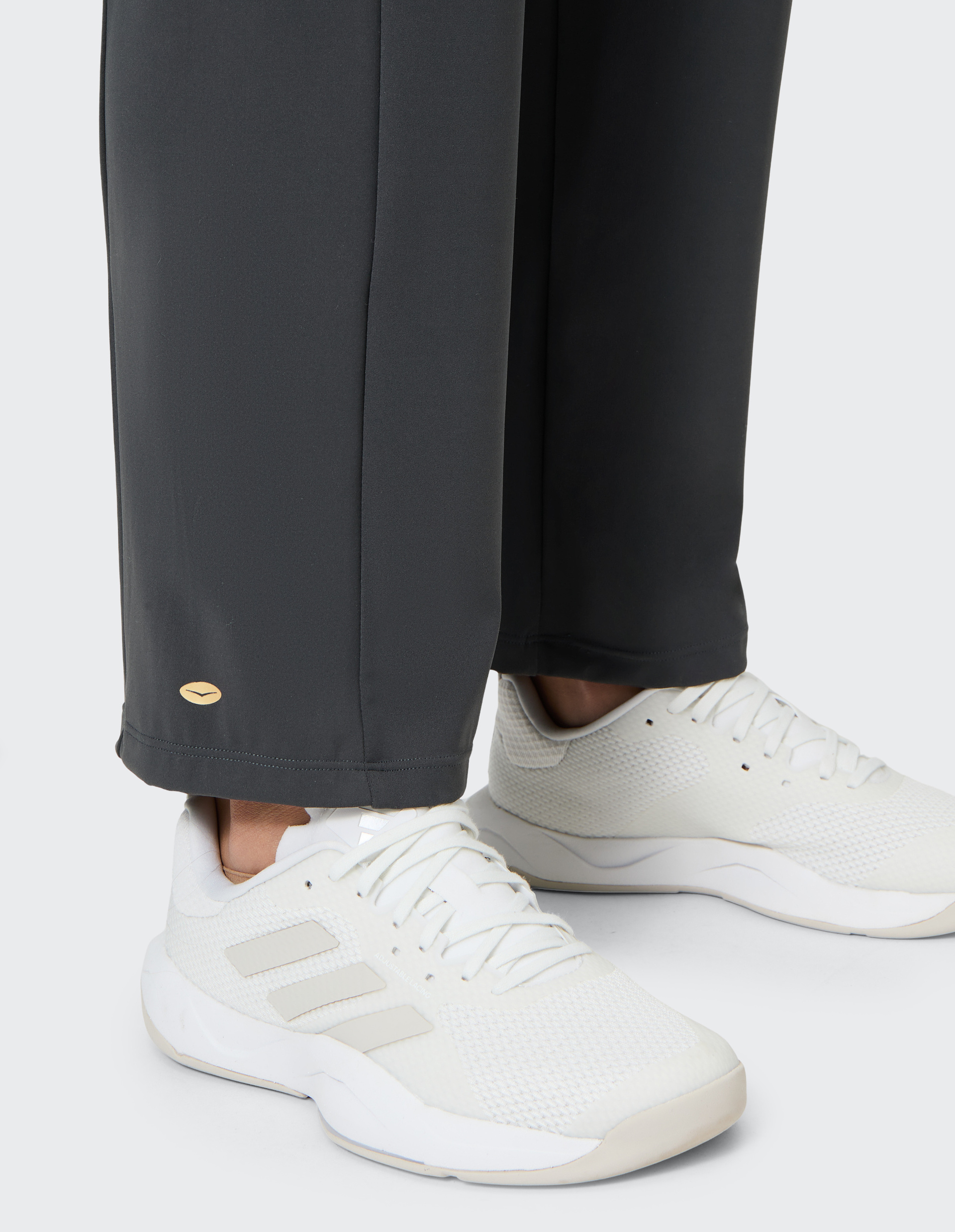 Weiße Sneaker mit parallelen Streifen, getragen zu dunkelgrauer Hose mit kleinem, ovalem, goldenem Emblem am Hosensaum.