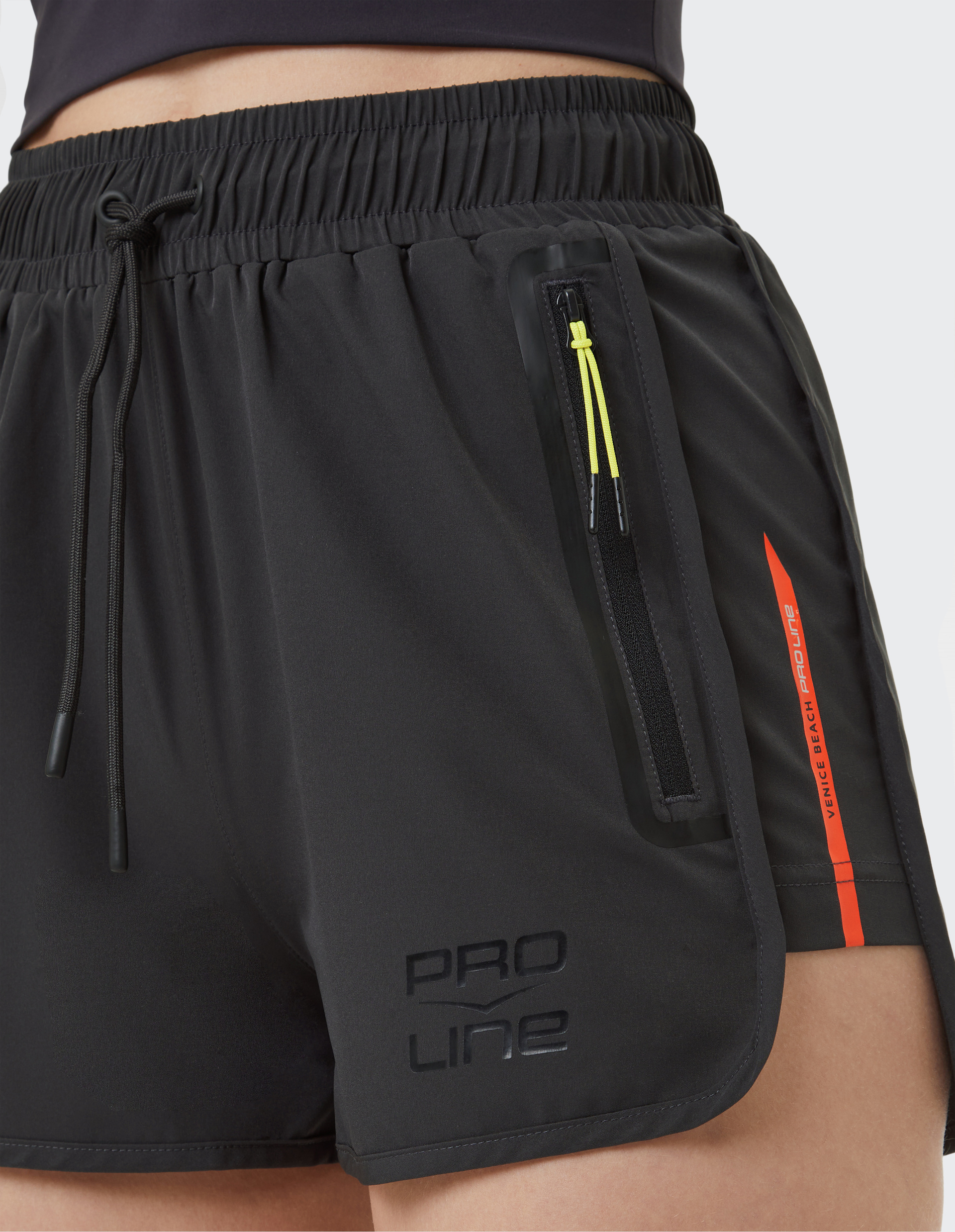 Nahaufnahme von schwarzen Sportshorts mit elastischem Bund, gelbem Zierband am Reißverschluss und rotem Streifen mit Schriftzug.