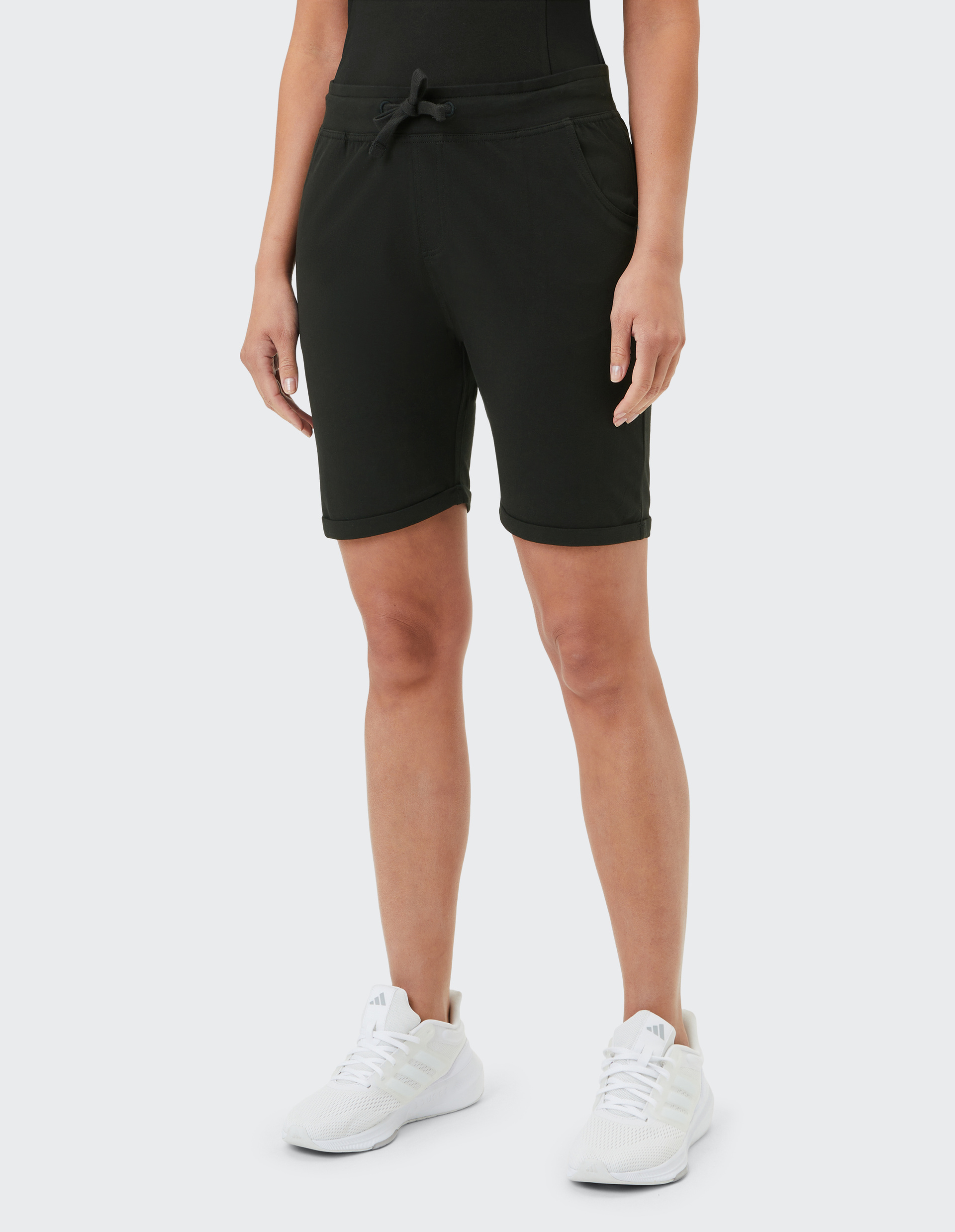 Person trägt schwarze Shorts mit Bindeband und Taschen sowie weiße Sneakers auf hellem Hintergrund, ab Hüfte abwärts gezeigt.