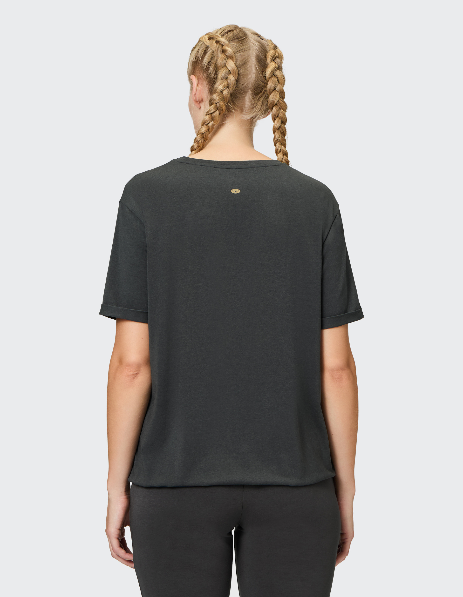 Eine Person mit blonden Zöpfen trägt ein dunkles T-Shirt und Hose, vom Rücken aus gesehen, vor einem grauen Hintergrund.