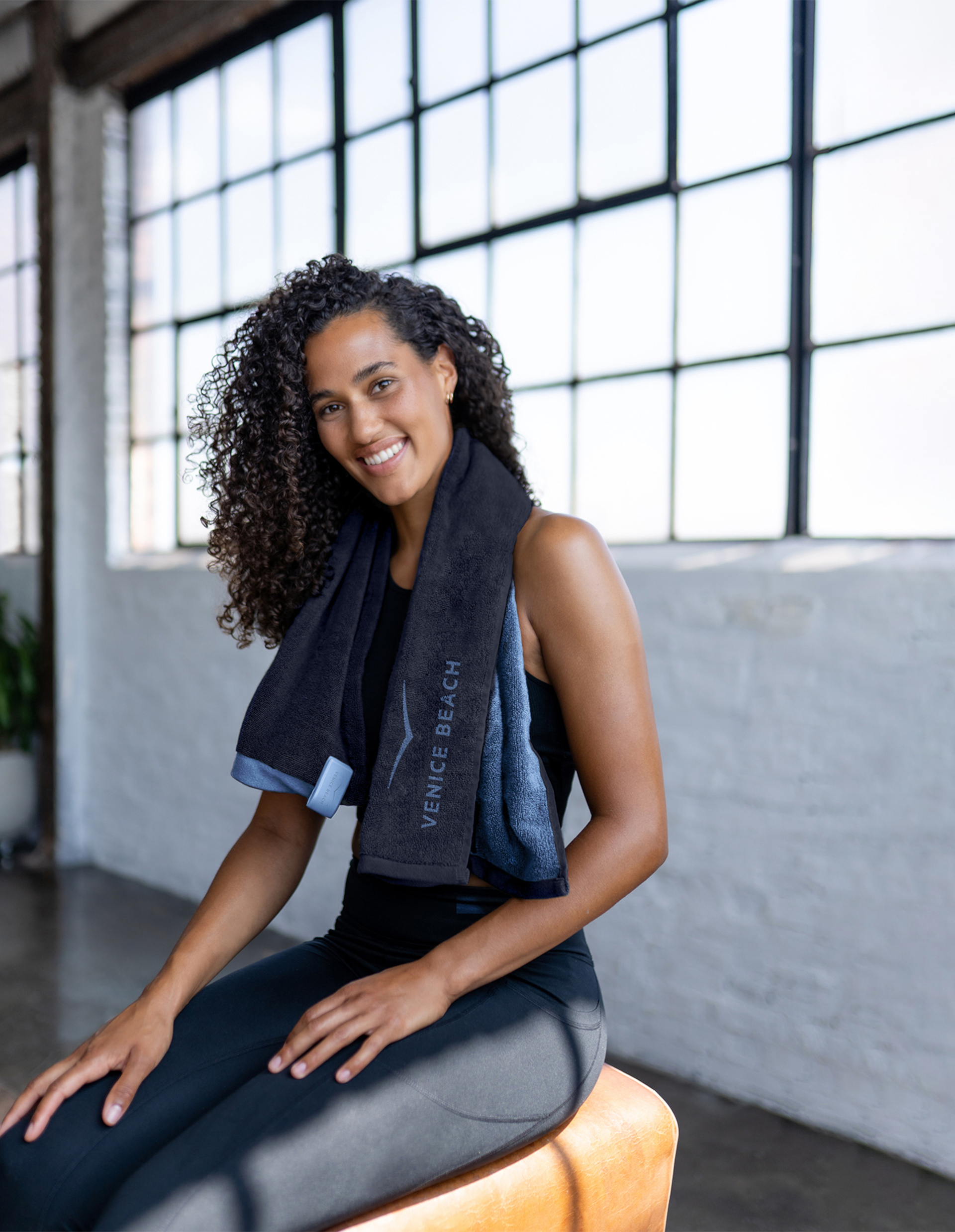 Eine Frau mit lockigem Haar sitzt in sportlicher Kleidung auf einem Lederwürfel, ein Handtuch um den Hals, vor großen Fenstern.