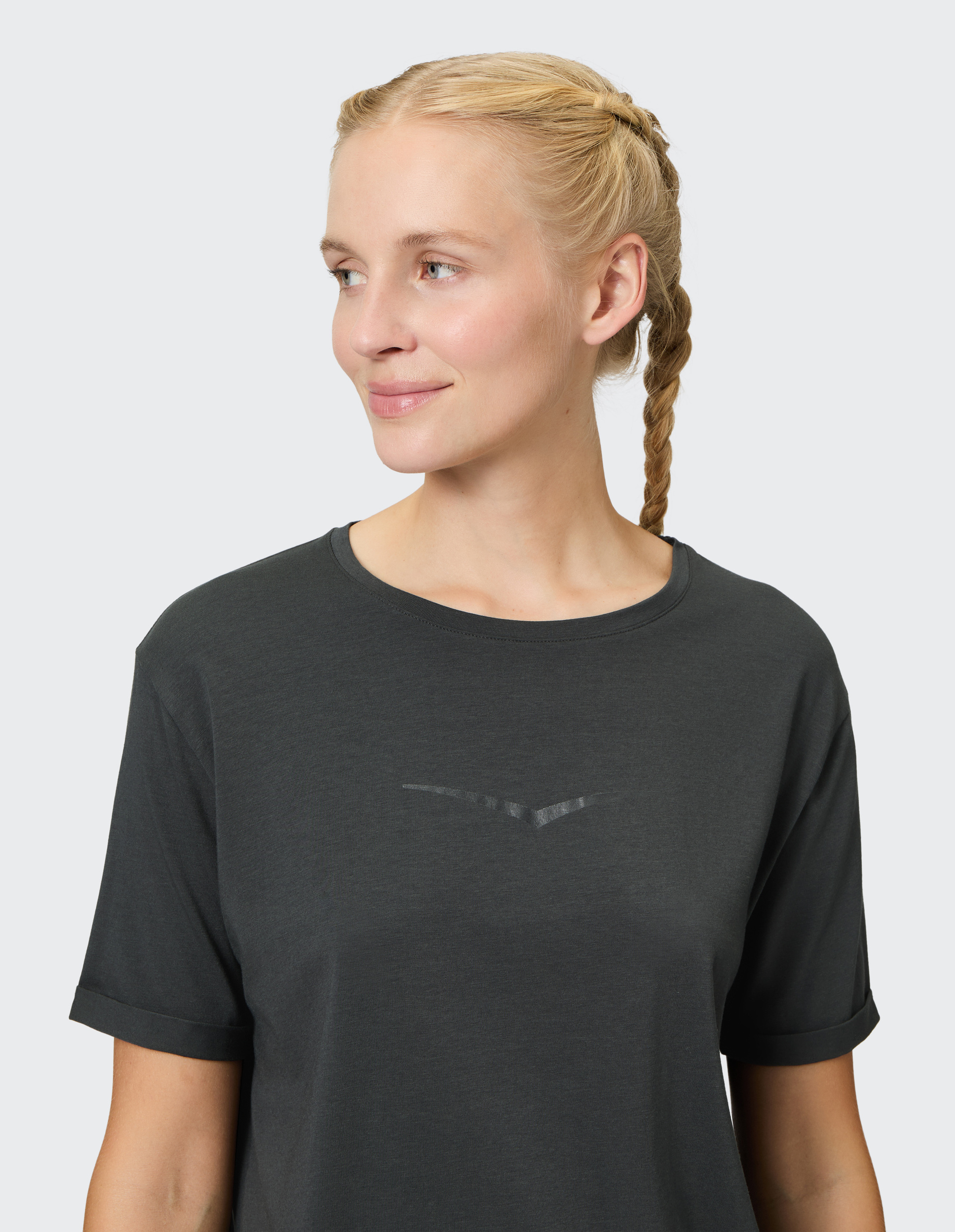 Eine Person mit geflochtenem Haar trägt ein dunkelgraues T-Shirt, blickt lächelnd nach links vor einem hellen Hintergrund.