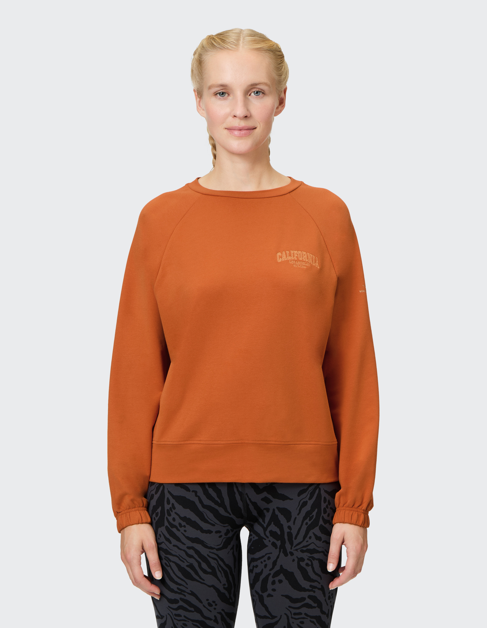 Eine lächelnde Person trägt einen orangefarbenen Pullover mit "California"-Aufdruck und schwarz-gemusterte Leggings vor grauem Hintergrund.