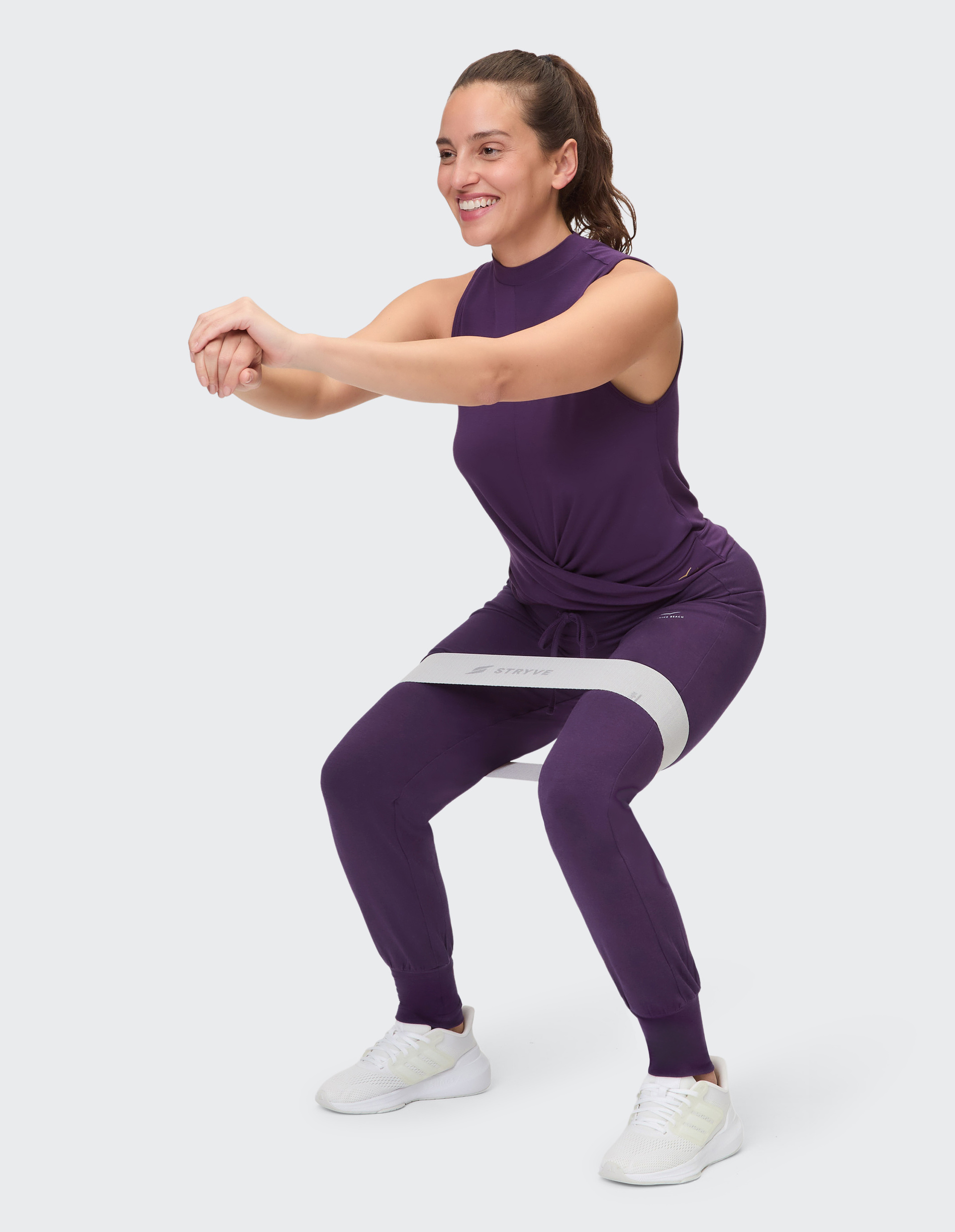 Frau in lila ärmellosem Oberteil und Jogginghose steht frontal, trägt weiße Sneaker und schaut direkt in die Kamera.