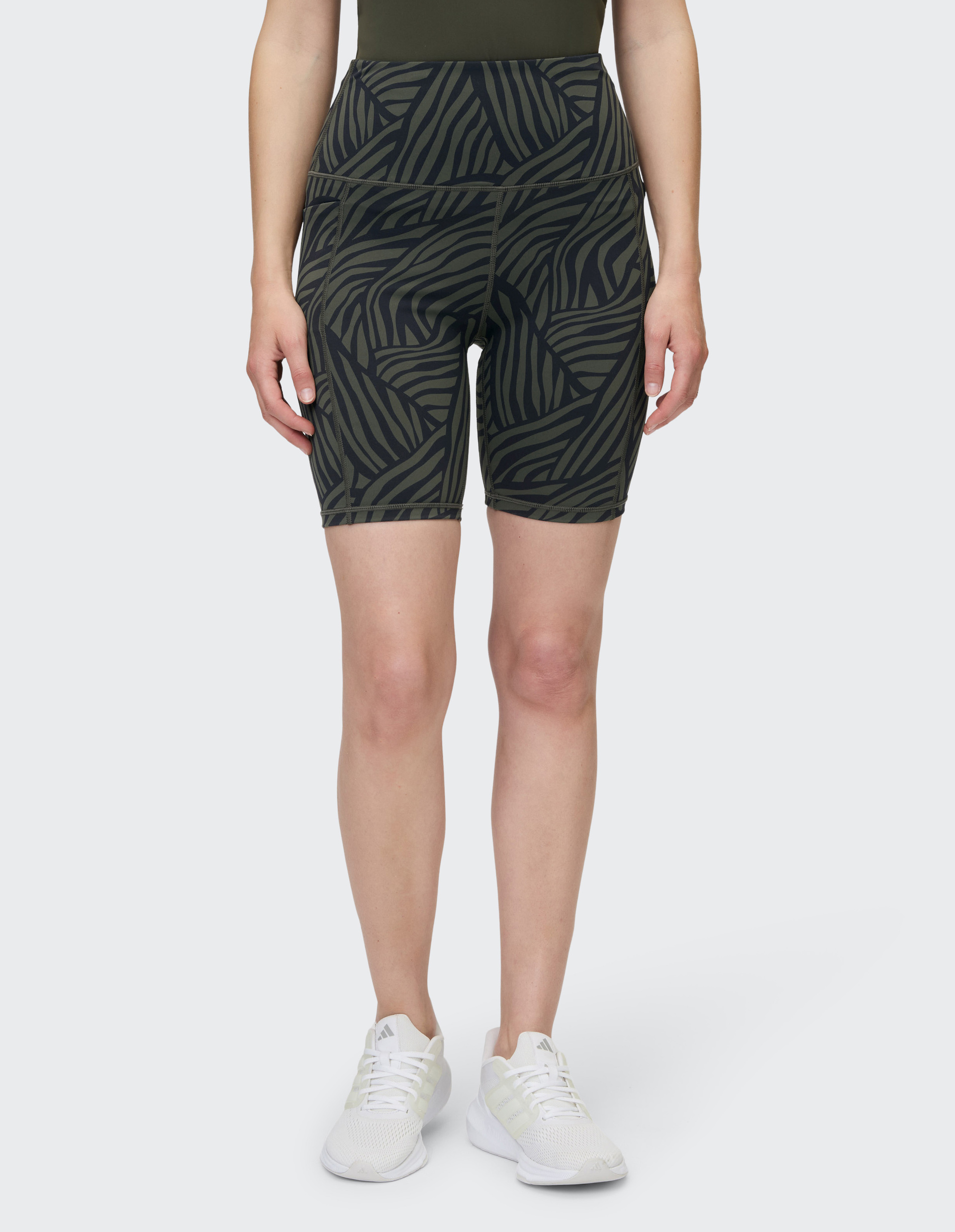 Frau trägt gemusterte, knielange Shorts mit Zebrastreifen in Schwarz und Oliv und weiße Sneaker, vor hellem Hintergrund.