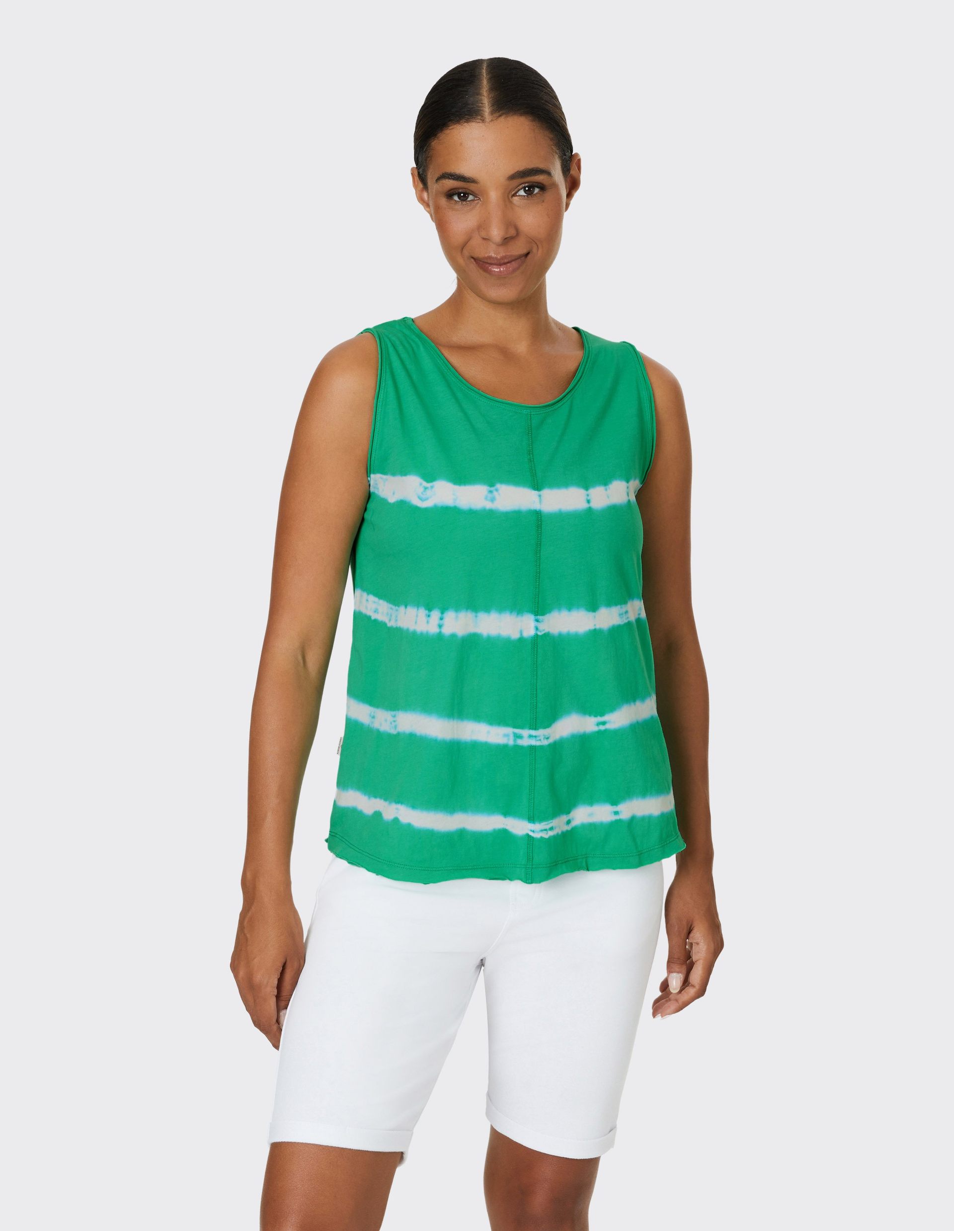 Frau trägt ärmelloses, grün-weiß gestreiftes Oberteil und weiße Shorts, lächelt leicht, vor hellem Hintergrund.