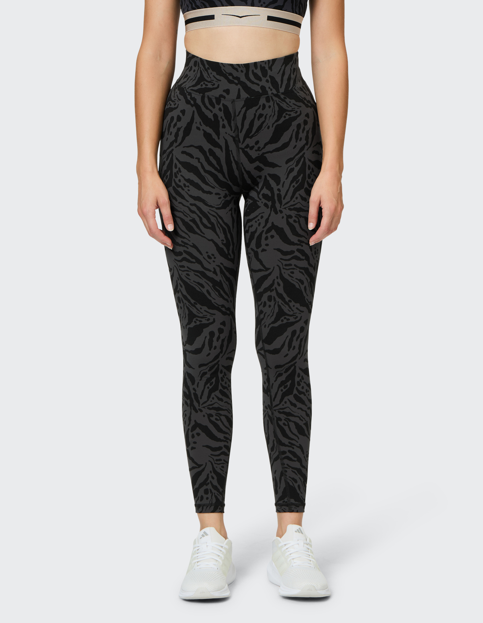 Frau trägt ein modernes Fitness-Set mit schwarzem, wild gemustertem Leggings und passendem Sport-BH, dazu weiße Sneaker.