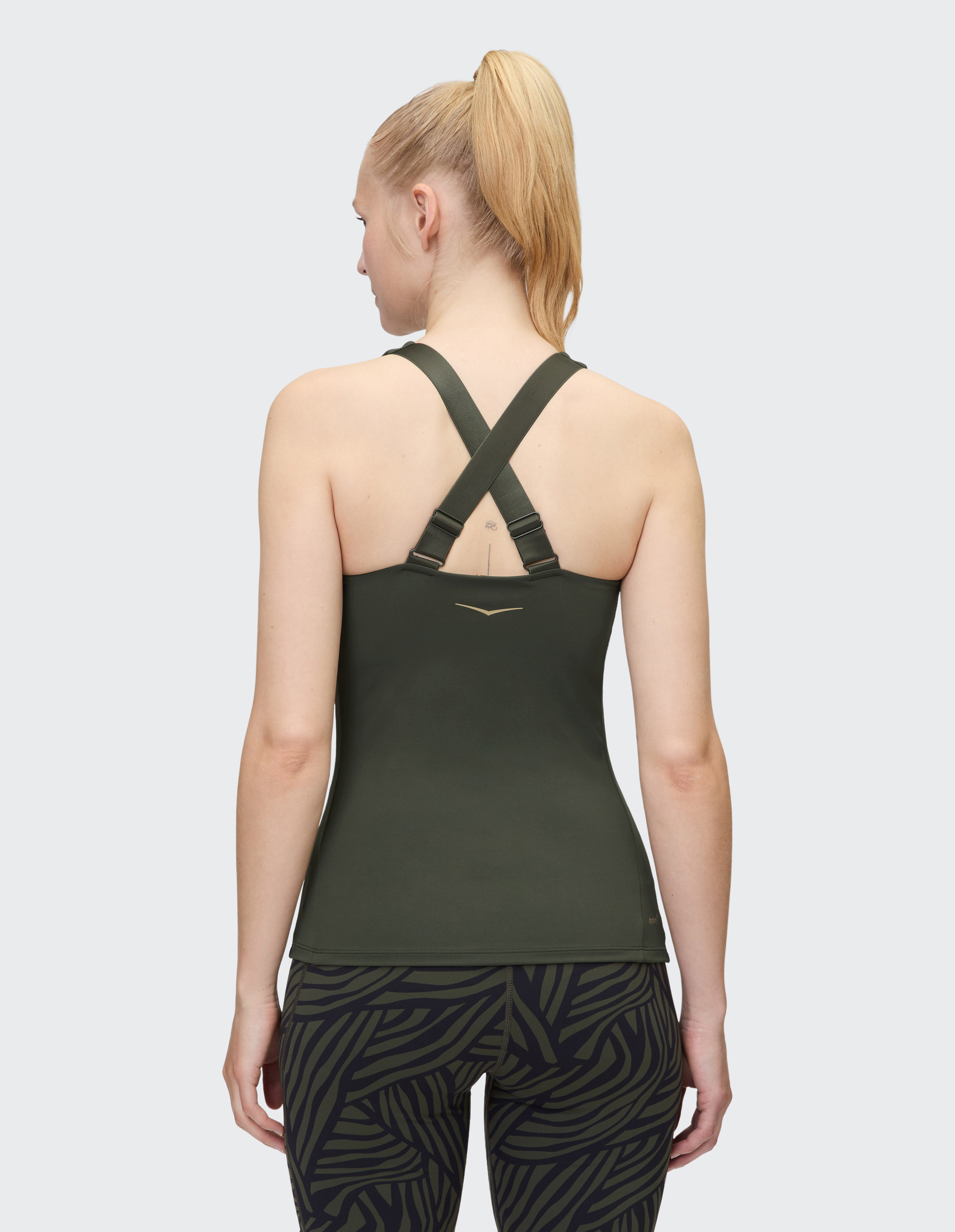 Eine Frau mit blondem Pferdeschwanz trägt ein olivgrünes Sporttop mit überkreuzten Trägern und gemusterten Leggings, von hinten gesehen.