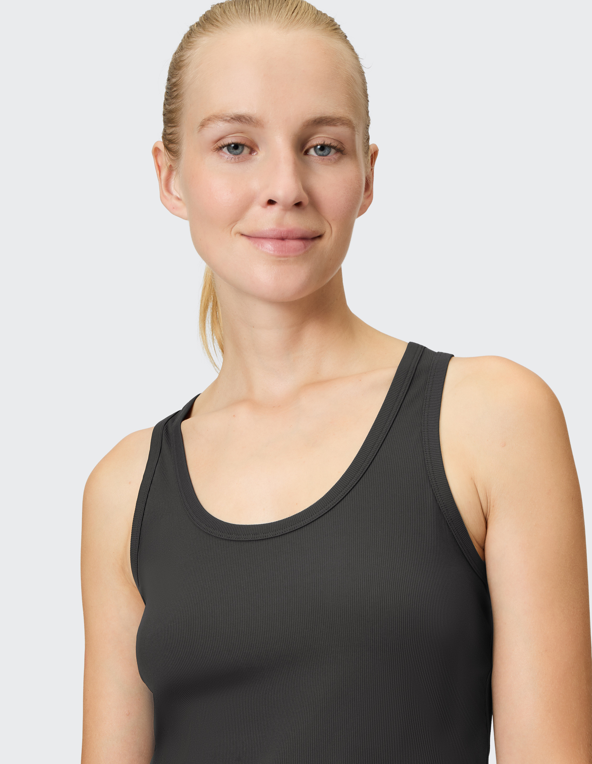 Eine Frau mit blondem Haar, lächelt leicht, trägt ein dunkles Tanktop und steht vor einem hellgrauen Hintergrund.