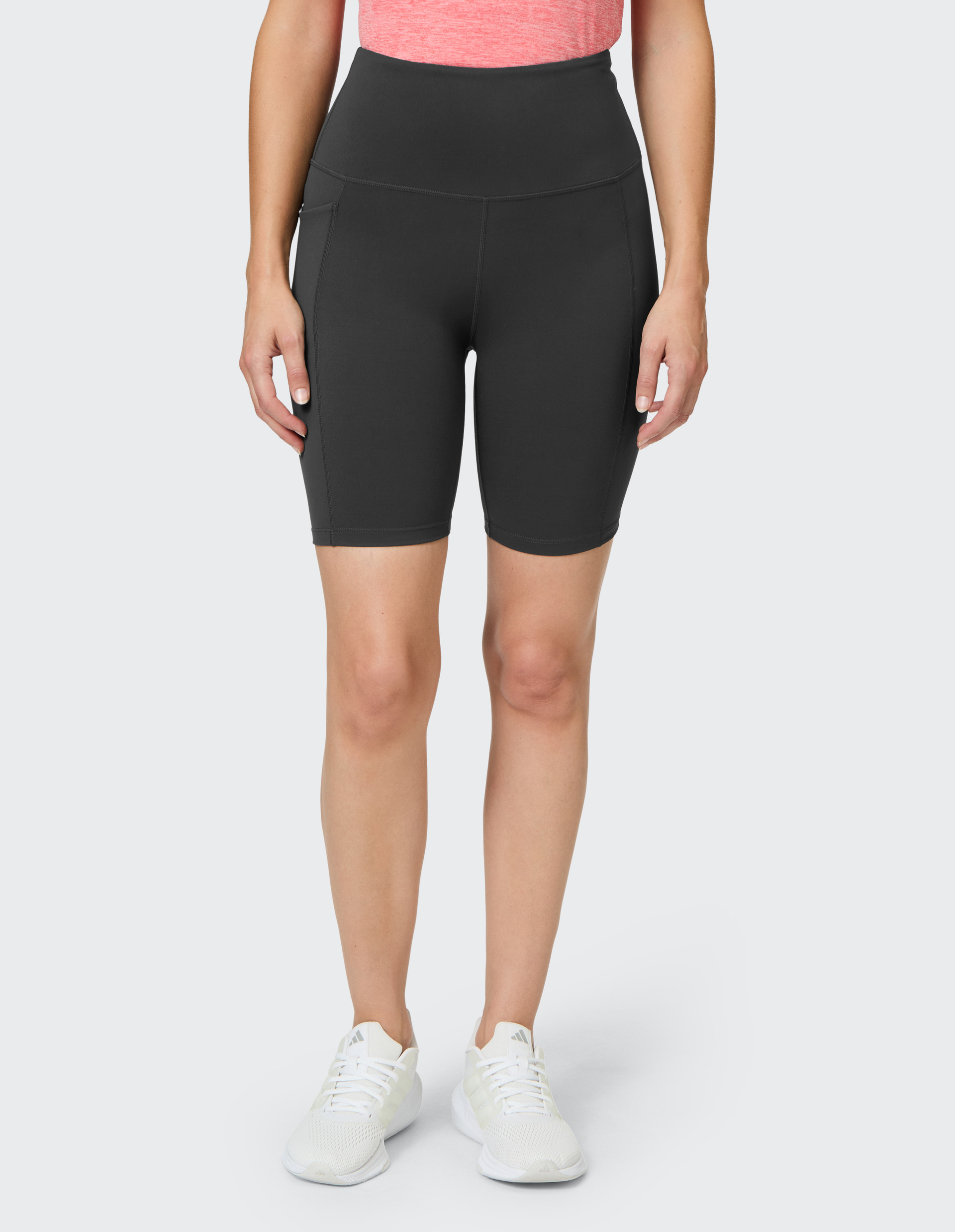 Frau trägt kurze, schwarze Sportleggings mit hohem Bund und Seitentaschen, kombiniert mit rosa Oberteil und weißen Turnschuhen.