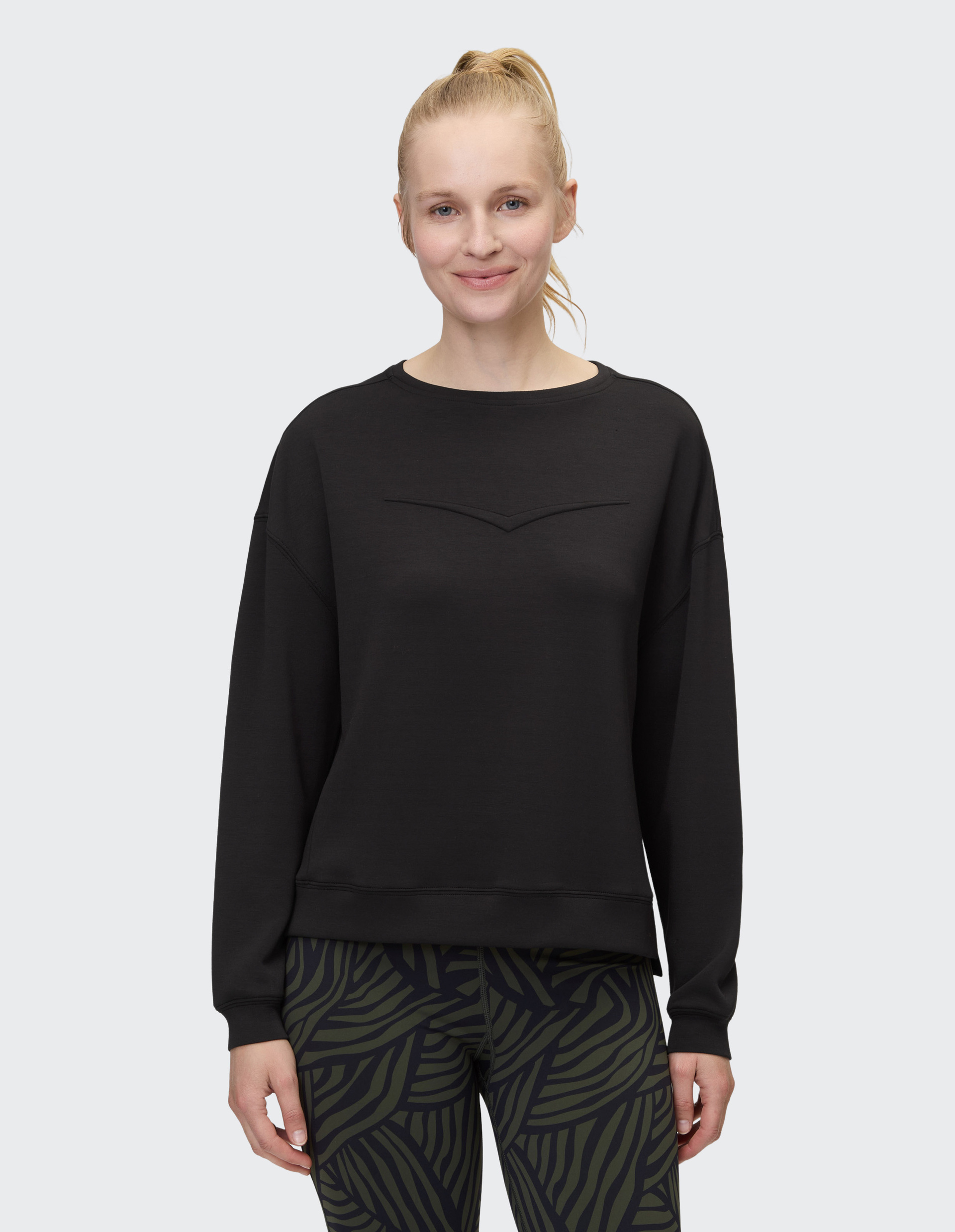 Person in schwarzem Pullover, gemusterten Leggings und blondem Haar im Pferdeschwanz vor neutralem Hintergrund lächelnd.