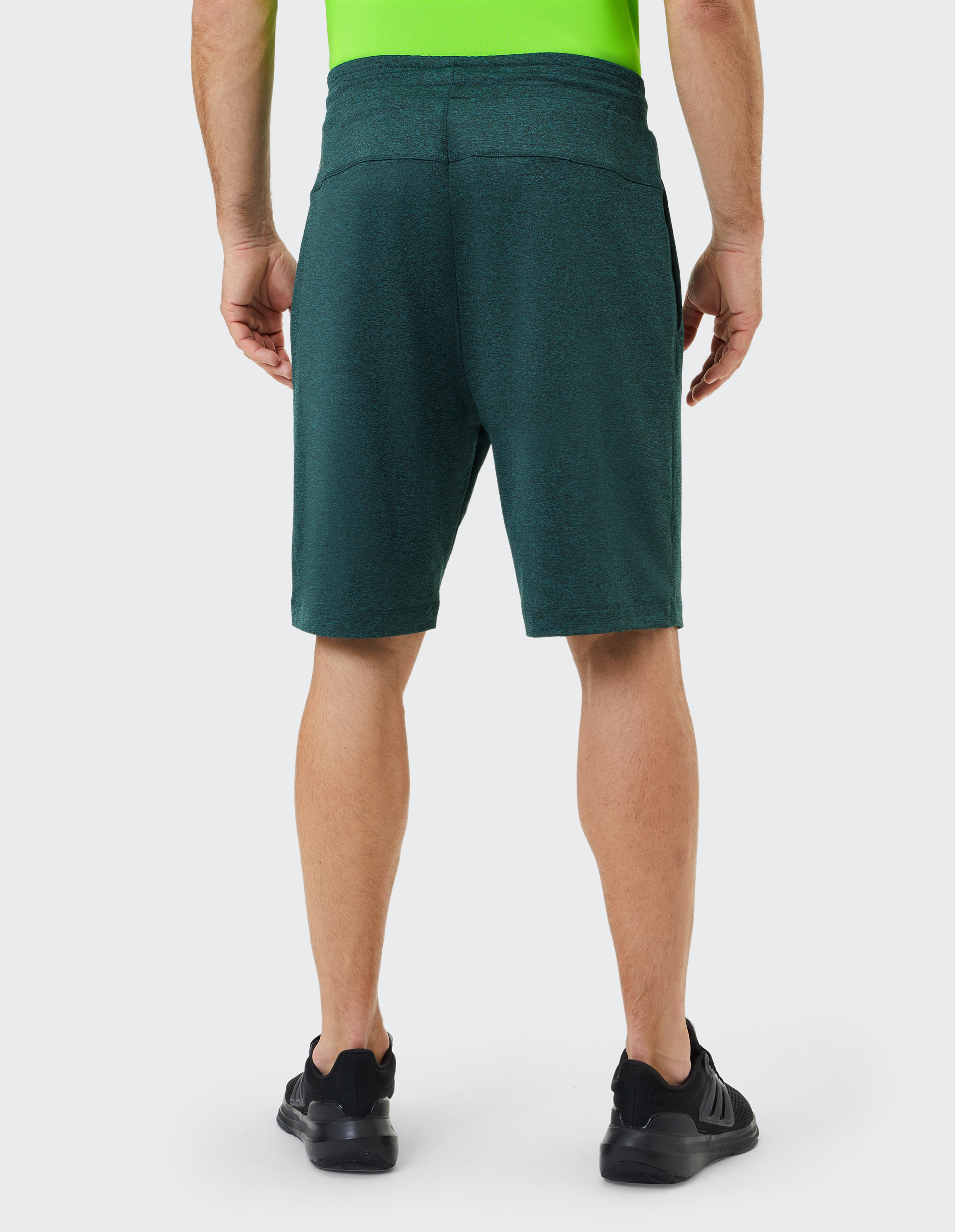 Eine Person trägt grüne Shorts und ein neonfarbenes Shirt, steht mit dem Rücken zur Kamera; schwarze Sneakers ergänzen das Outfit.