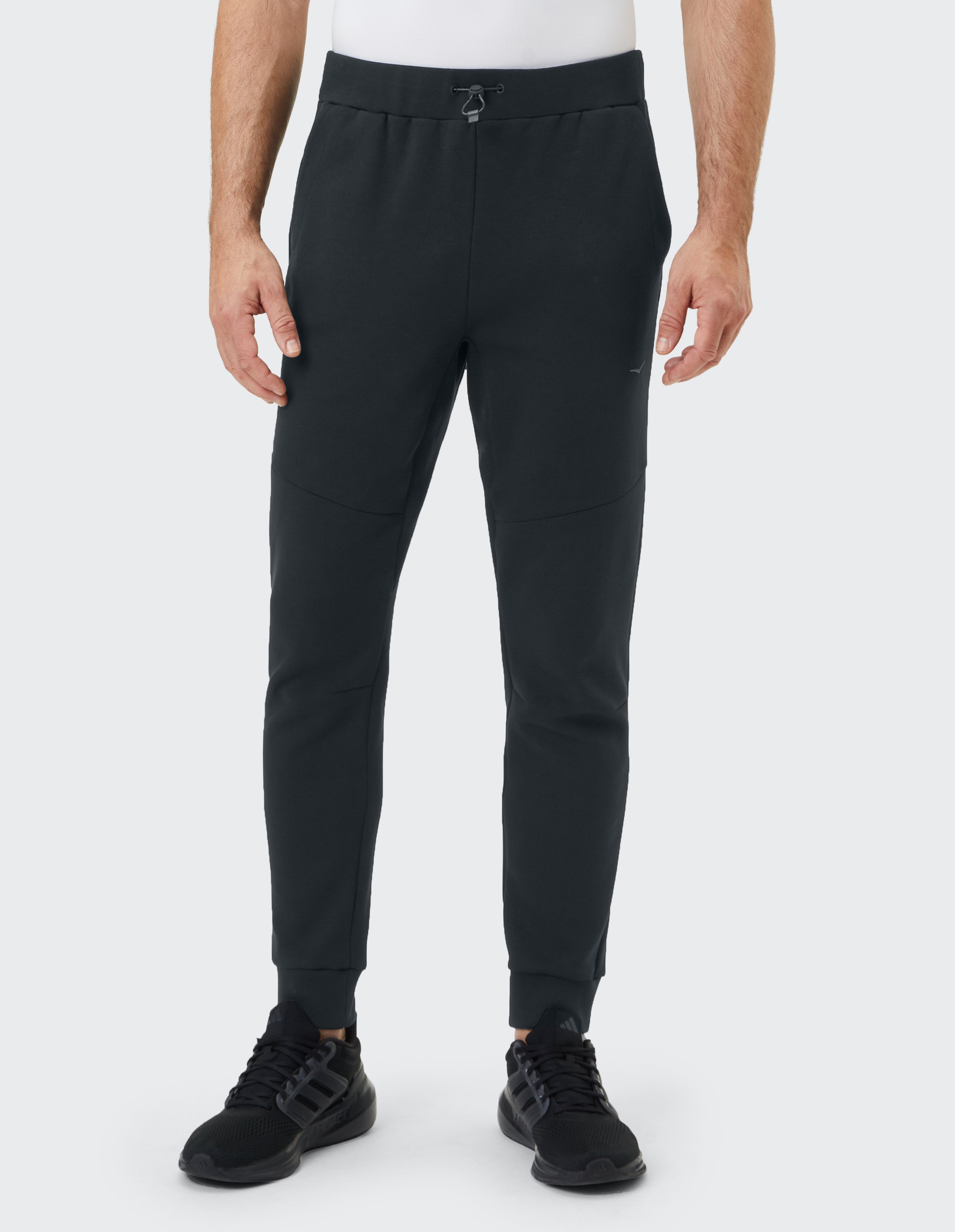 Eine Person trägt eine schwarze Jogginghose mit elastischem Bund und Zugband, dazu schwarze Sportschuhe auf grauem Hintergrund.