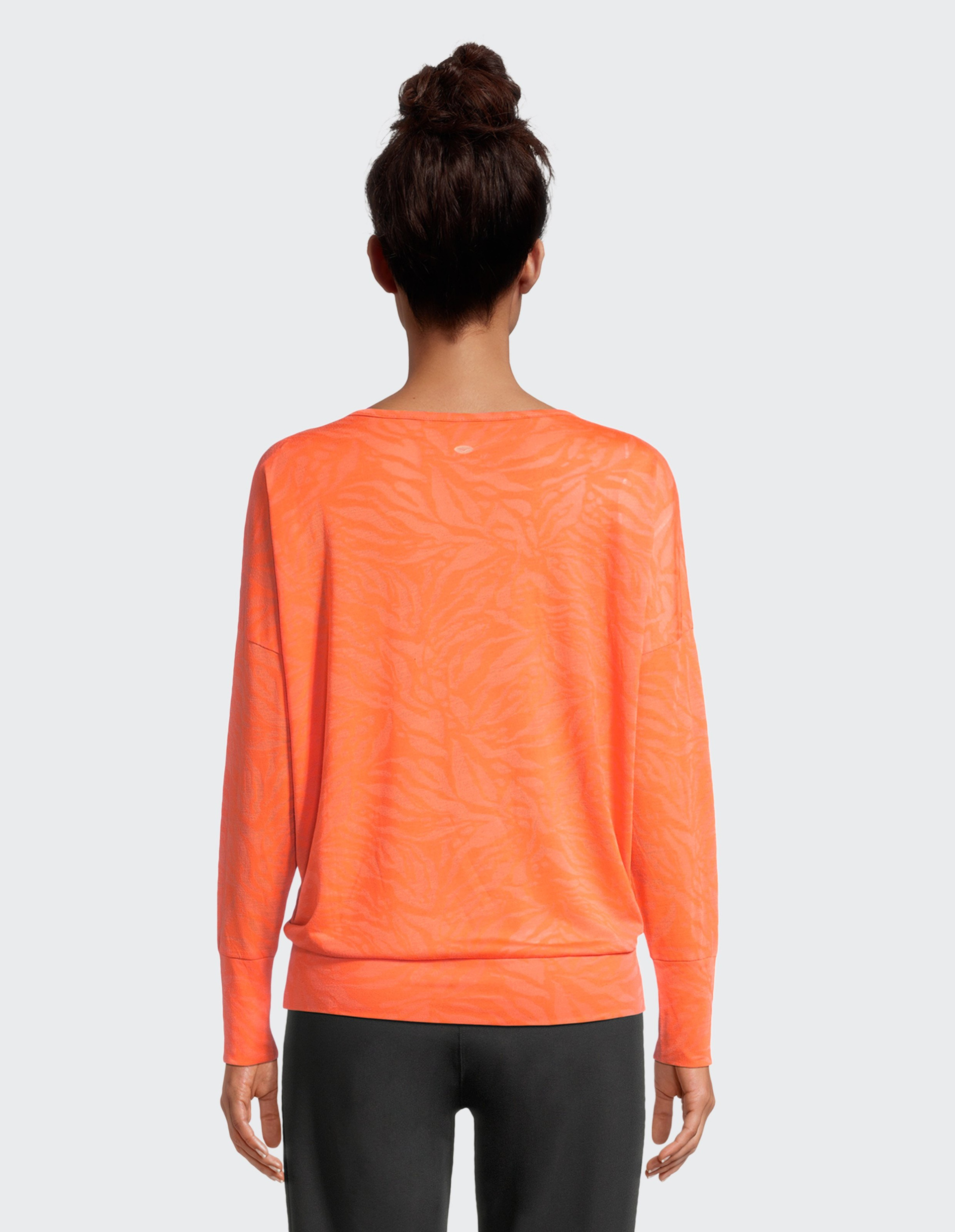 Person von hinten, trägt leuchtend oranges, langärmeliges Oberteil mit subtilen Mustern und dunkle Hose, Haare zu einem Dutt gebunden.