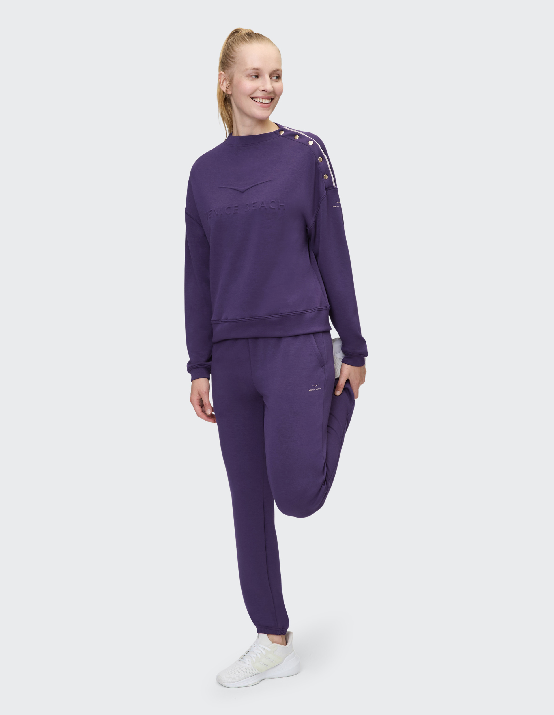 Person in lila Trainingsanzug mit Aufschrift "Venice Beach", weißem Hintergrund und weißen Sneakern, lächelt frontal in die Kamera.