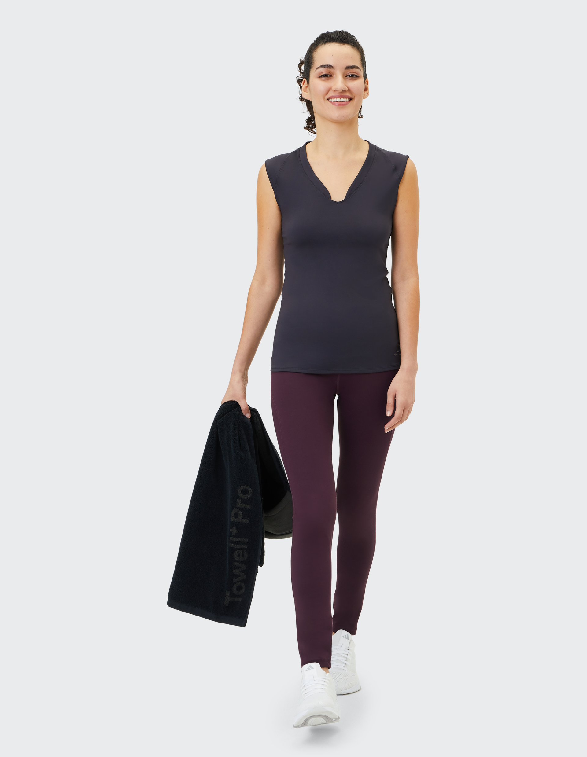 Frau in Sportkleidung, mit schwarzem Shirt und lila Leggings, hält ein schwarzes Handtuch und lächelt, geht nach rechts.