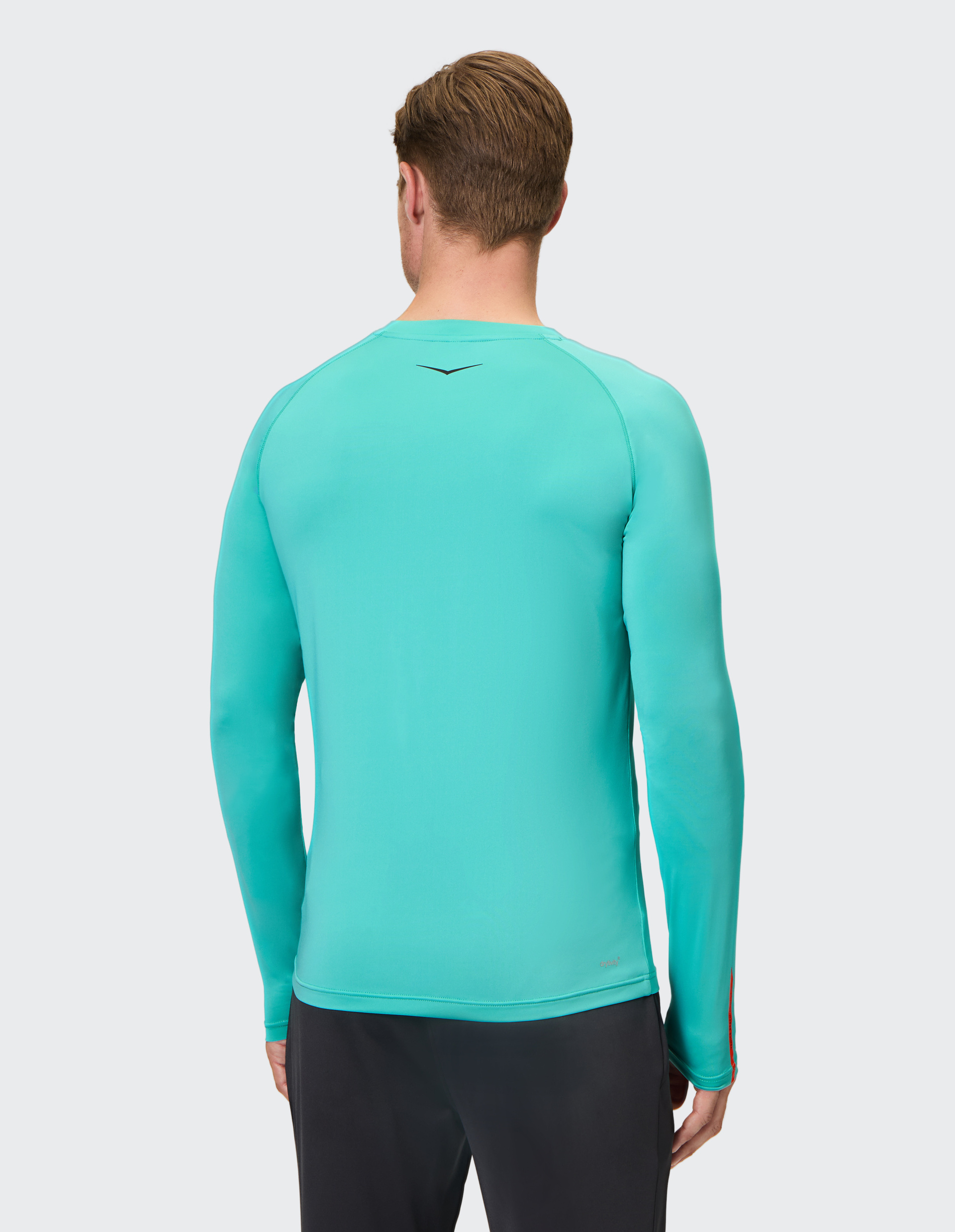 Ein Mann von hinten in einem hellblauen Langarmshirt und dunklen Hosen vor grauem Hintergrund.