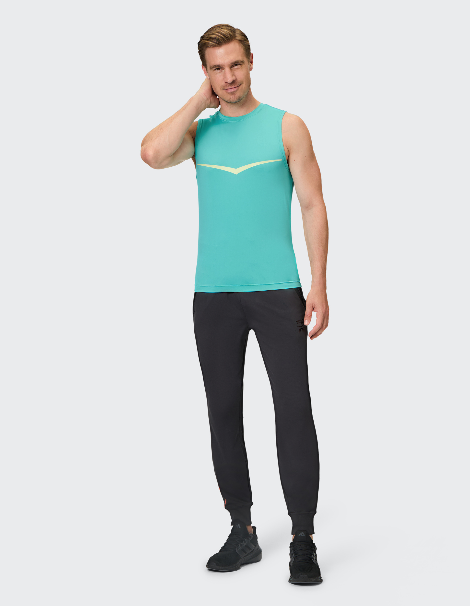 Junger Mann in türkisfarbenem, ärmellosem Shirt und schwarzer Sporthose, fasst sich lächelnd an den Nacken vor hellem Hintergrund.